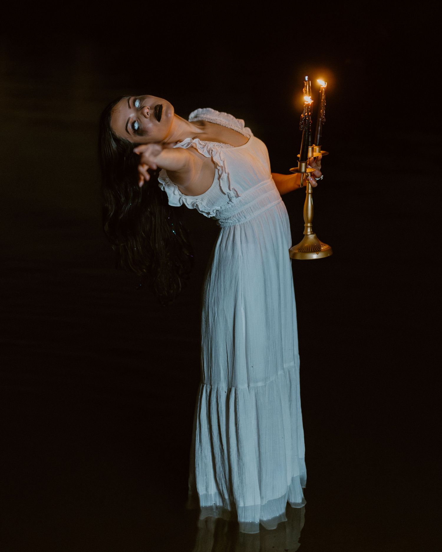 Figure in flowing light blue dress poses dramatically with candlelight in dark setting.