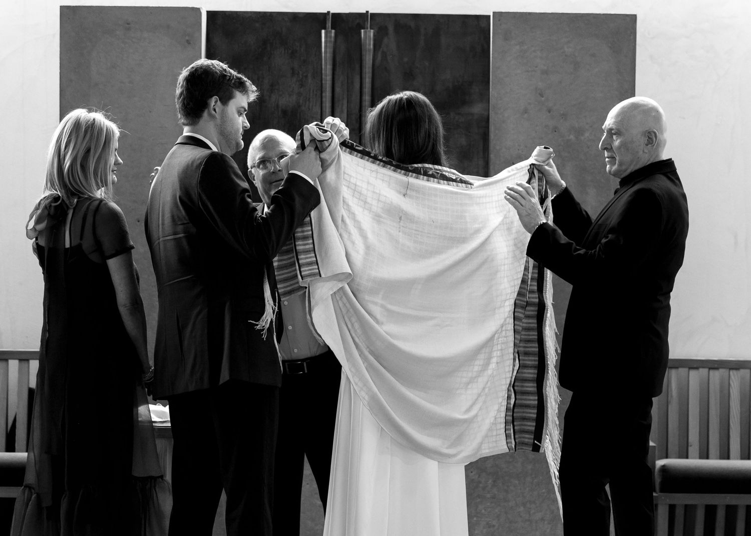 Black and white photo of a Jewish wedding ceremony with couple under tallit, officiant, and family members.