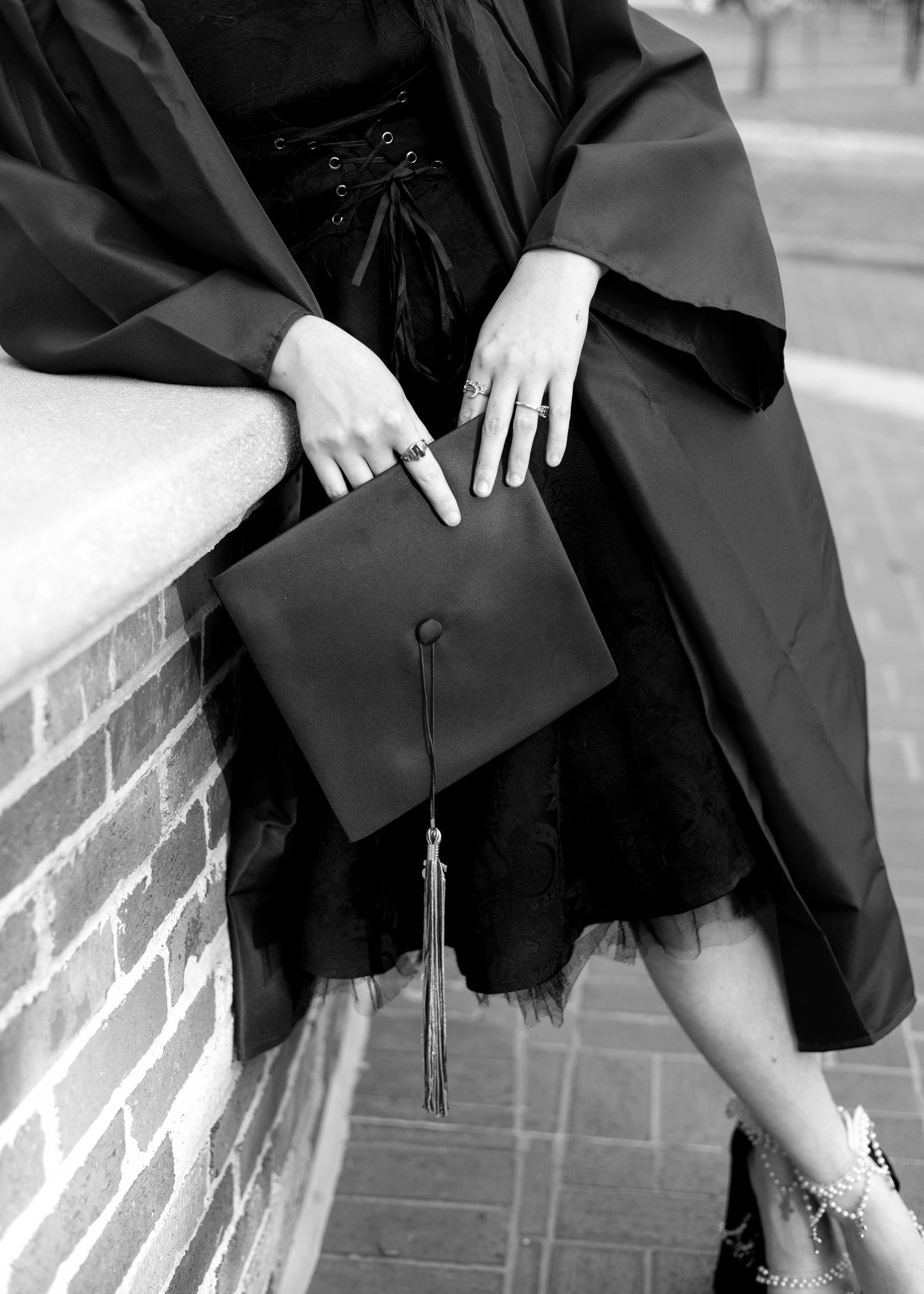 Black and white photo of a graduate in cap and gown holding a mortarboard, seated on a brick wall.