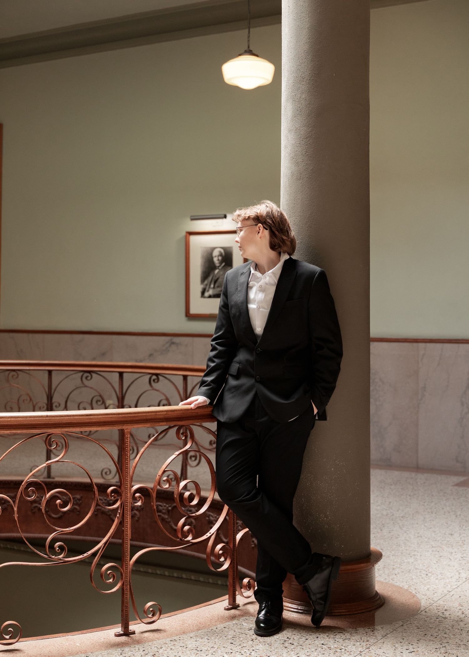 A man in a black suit leans against a column by ornate railings in an elegant interior with a pendant light.