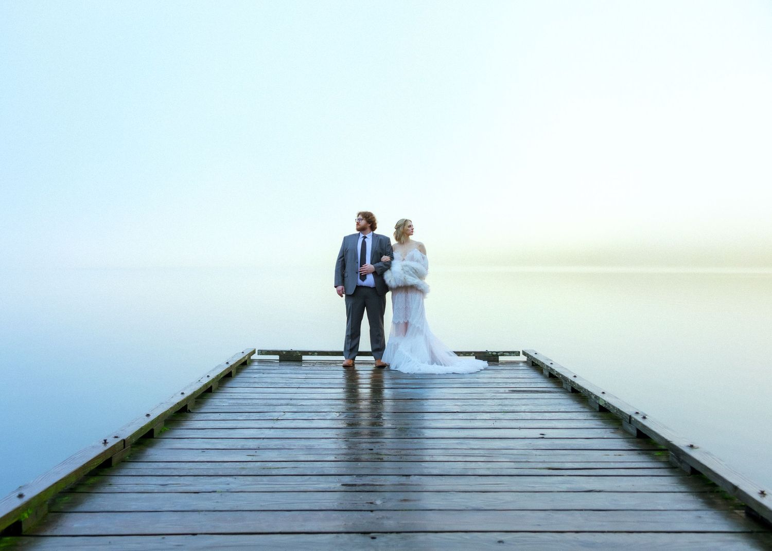 Bride and groom standing together on a wooden pier over calm misty water on their wedding day.