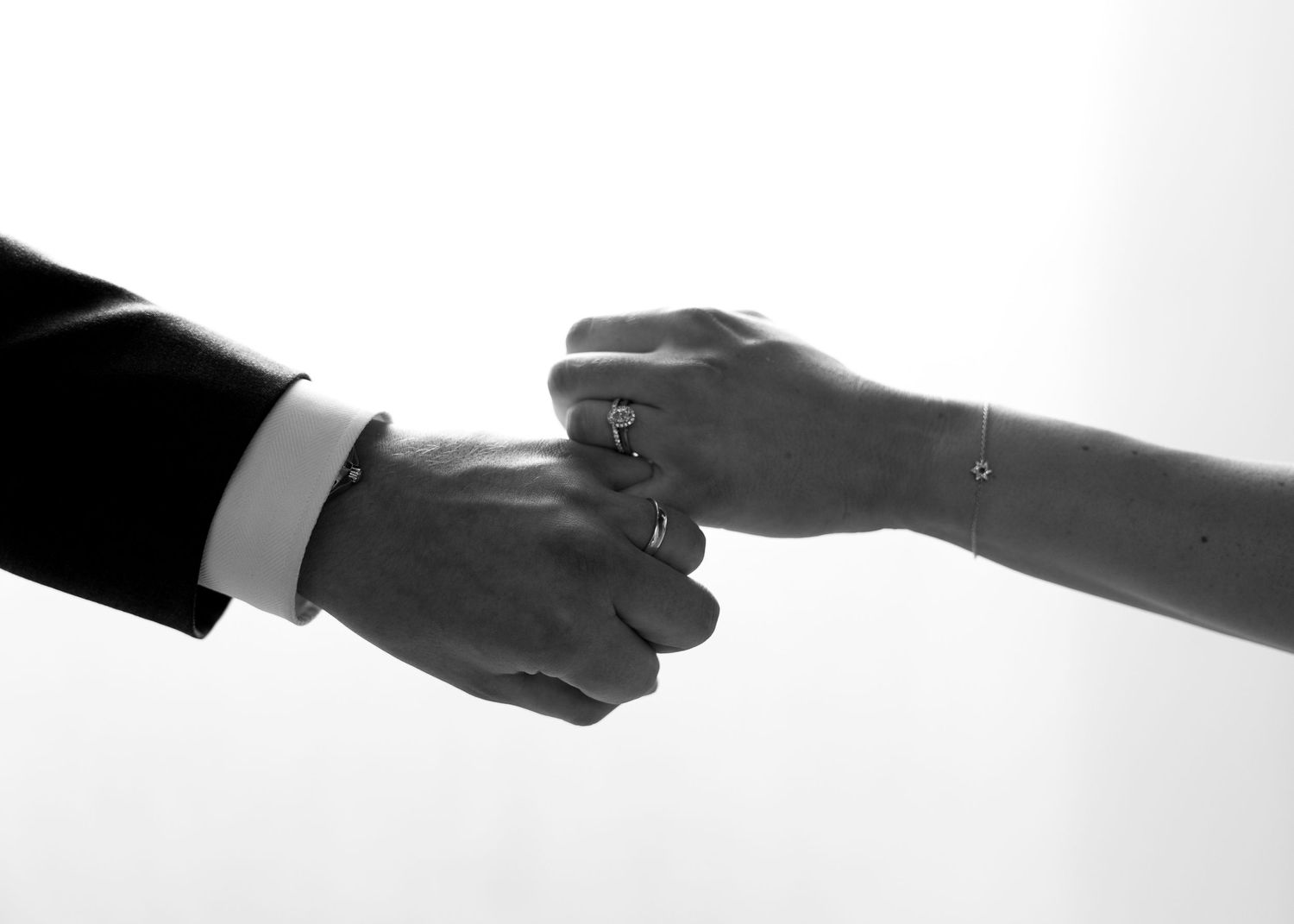 Black and white photo of a couple holding hands, showcasing a delicate bracelet and wedding ring.