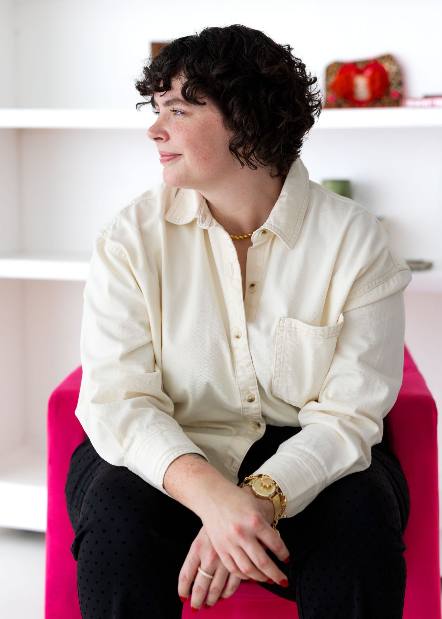 Person in cream colored button-down shirt sits casually on pink chair against white minimalist background.