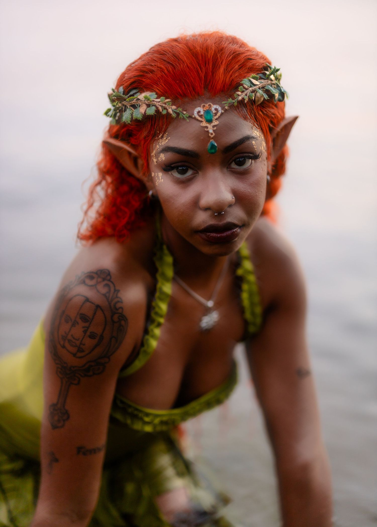 Fantasy-inspired photoshoot with vibrant red hair, nature elements and ornate accessories against ocean backdrop.