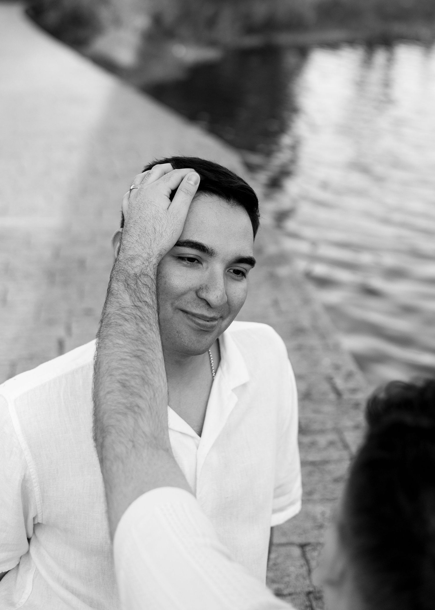 Black and white photo of a smiling man in white shirt touching his hair near a waterfront.