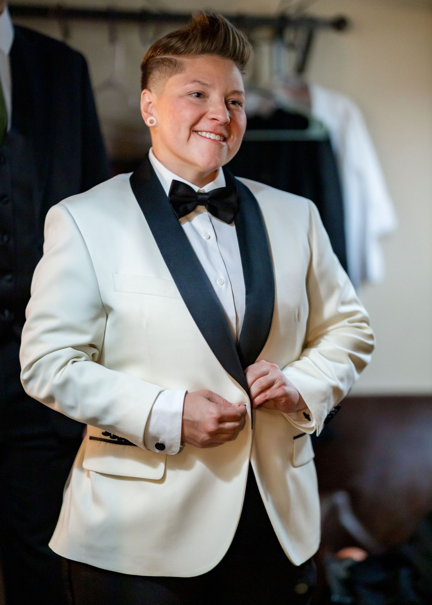 Person buttoning a cream tuxedo jacket with black lapels and bow tie at a wedding.