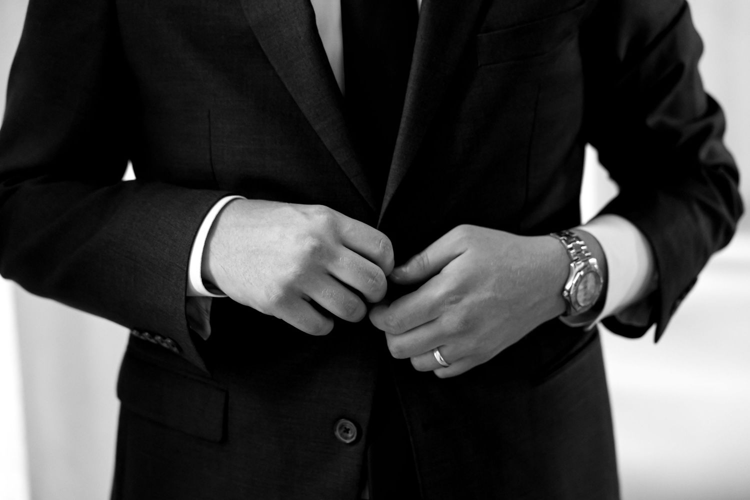Groom in a dark suit fastening jacket buttons with a luxury watch on his wrist in black and white photo.