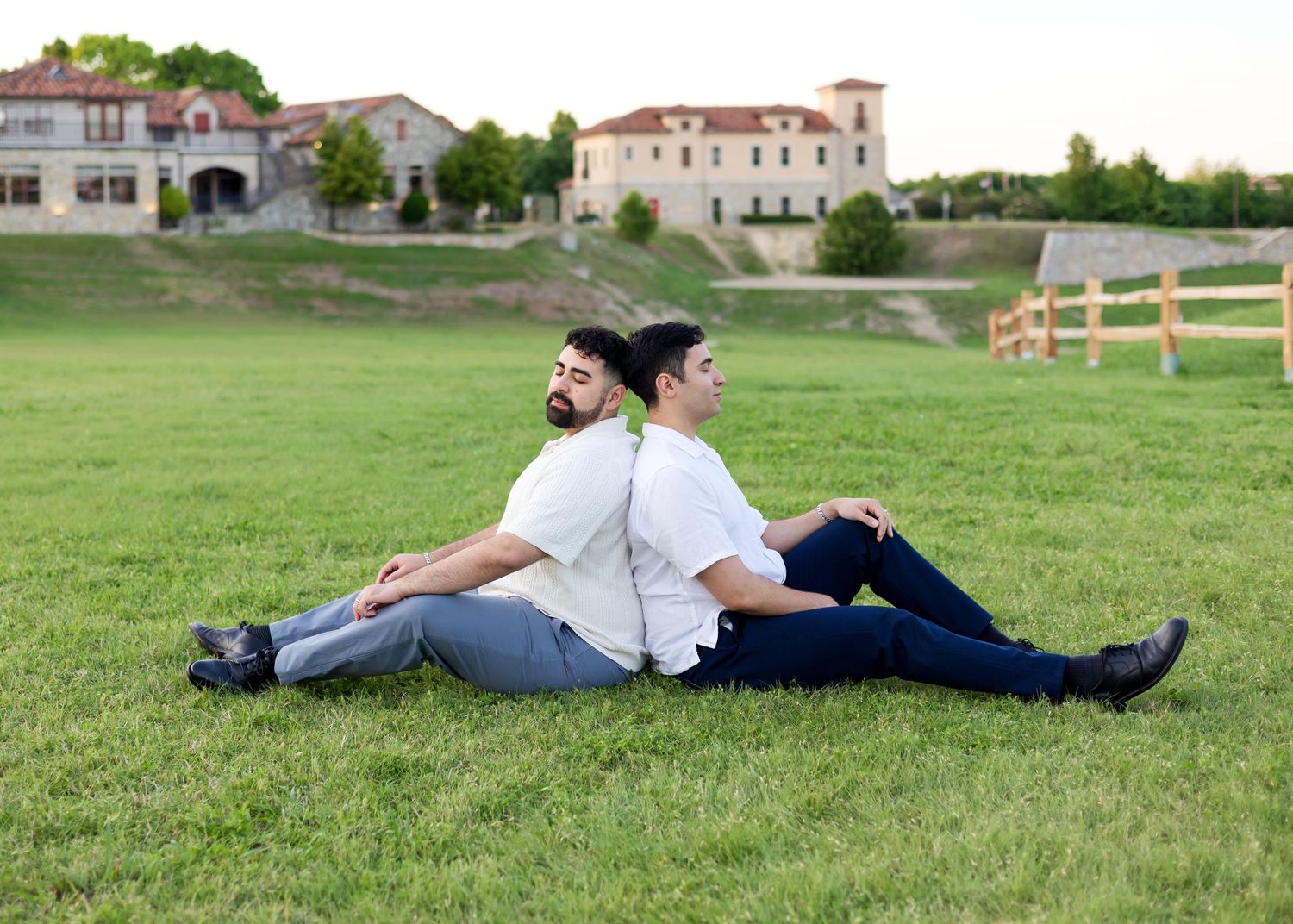 Two people sitting back-to-back on green grass with a grand estate building in the background.