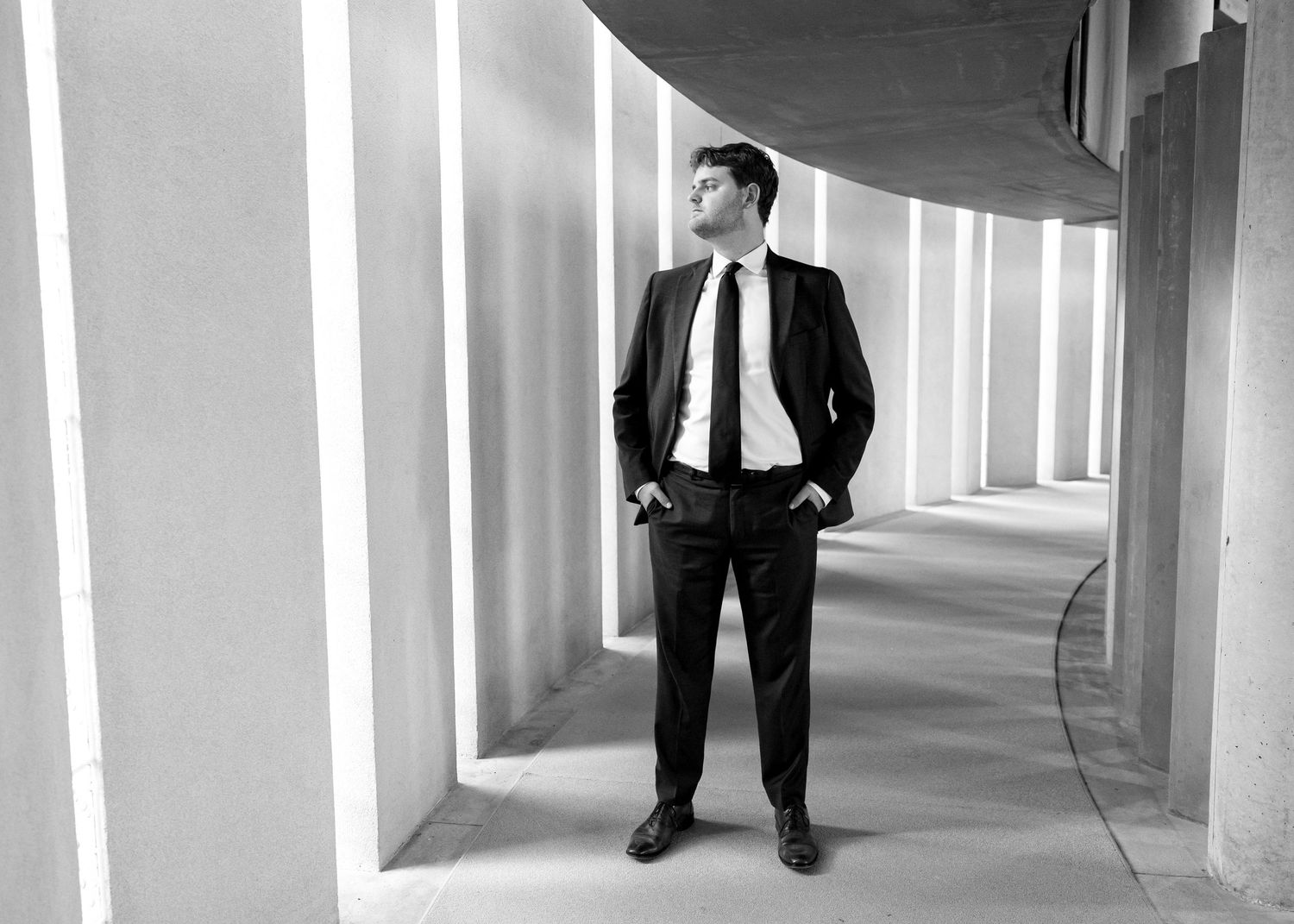 Dapper groom in black suit standing confidently in curved architectural corridor, black and white.
