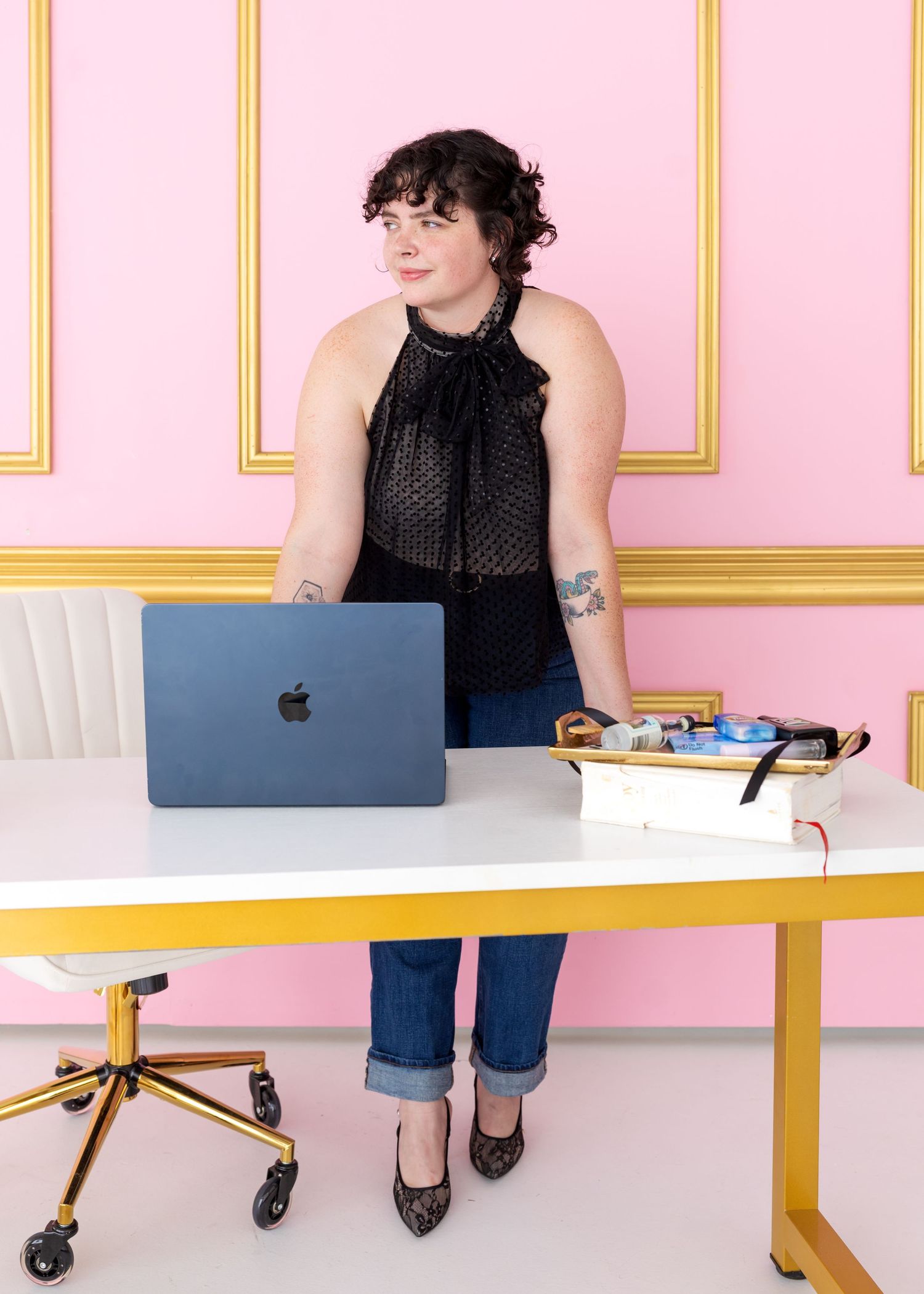 Person in black top works at modern desk setup with MacBook laptop against pink wall with gold trim details.