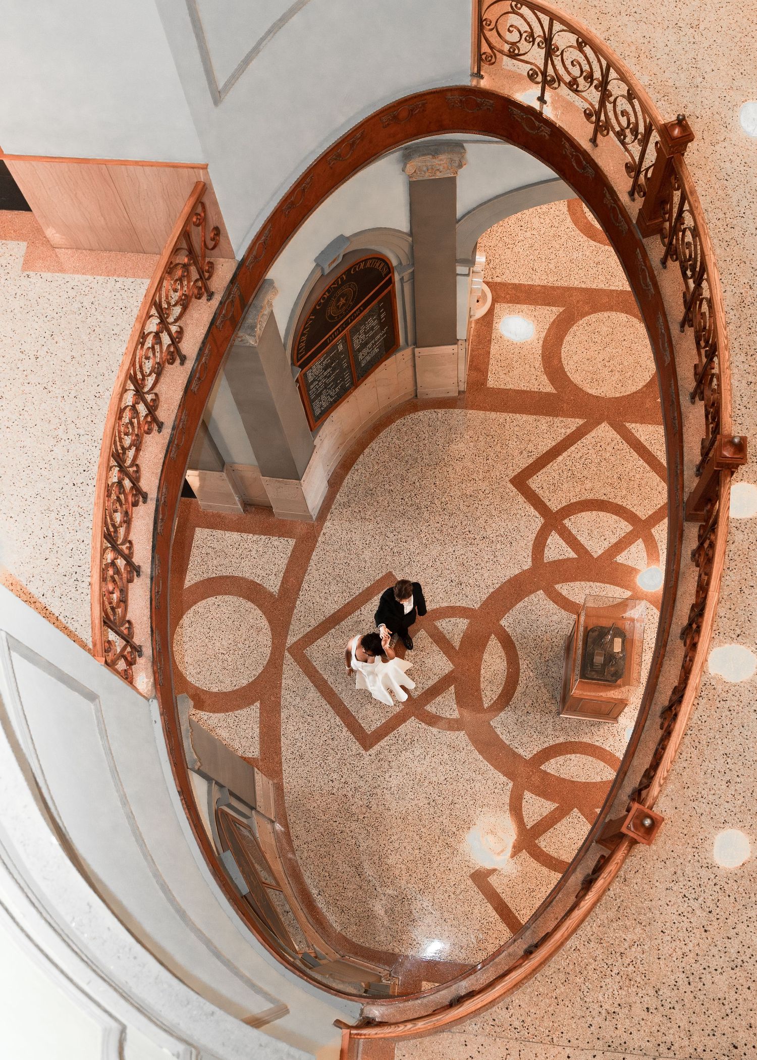 Aerial view of a couple embracing on ornate terrazzo floor beneath an elegant oval copper railing staircase.