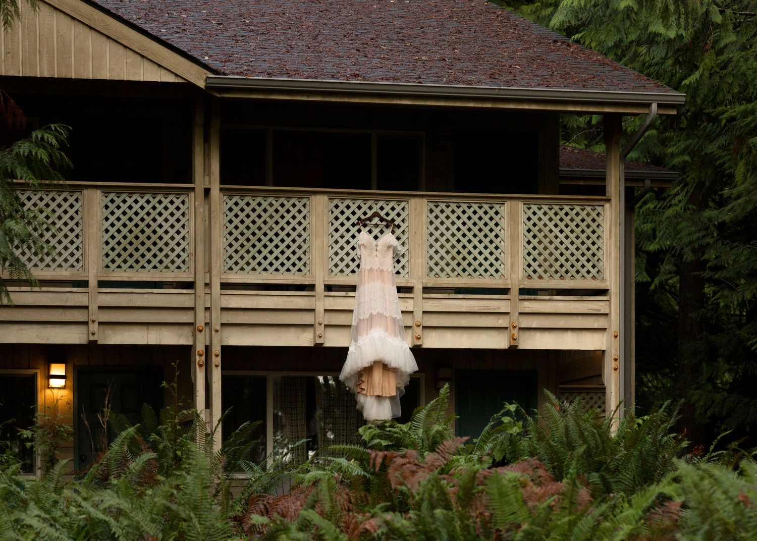 Wedding dress hanging on a wooden lodge balcony surrounded by lush green ferns and trees.