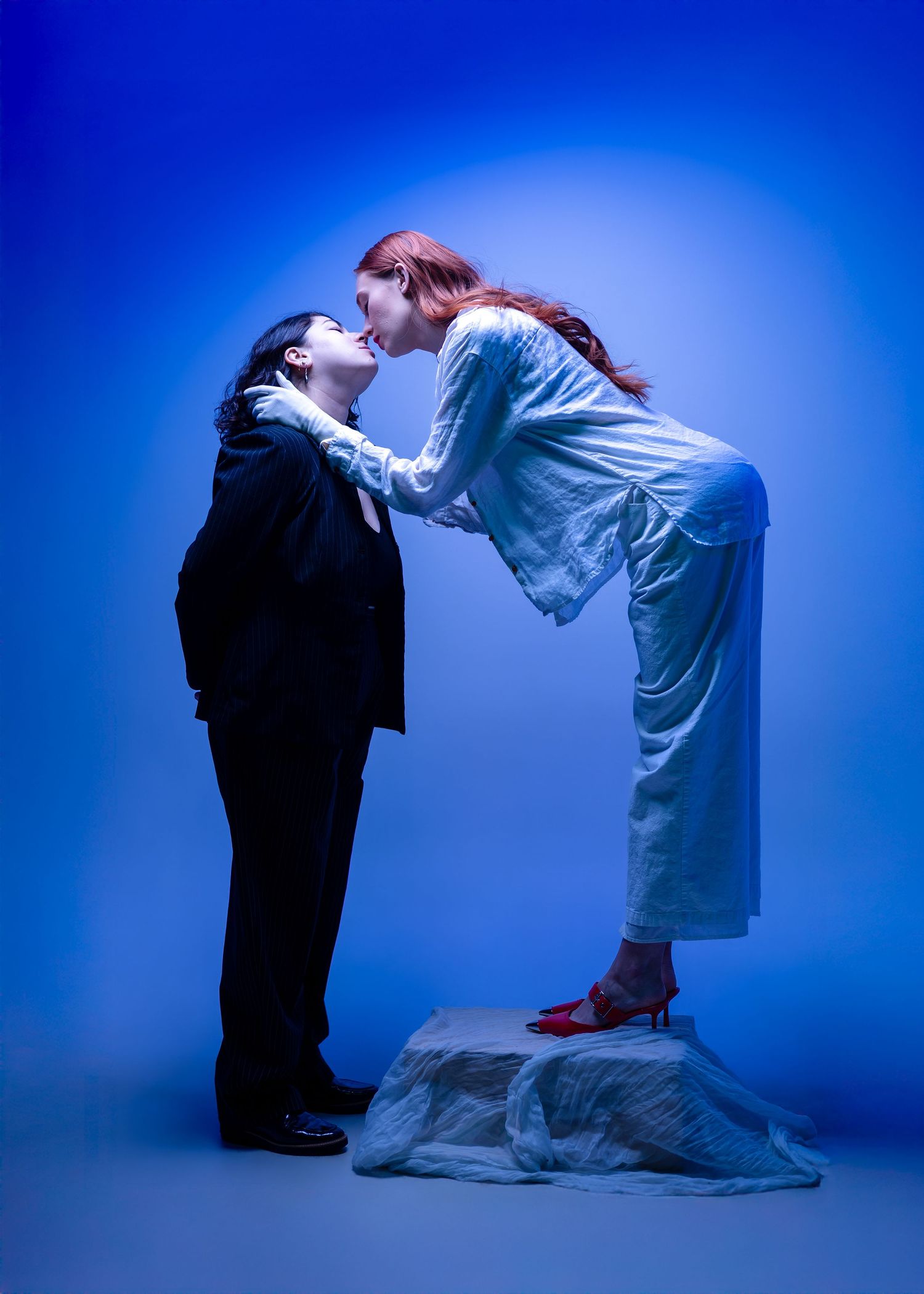 Dramatic studio portraits against blue backdrop showing interaction between two figures in contrasting outfits.