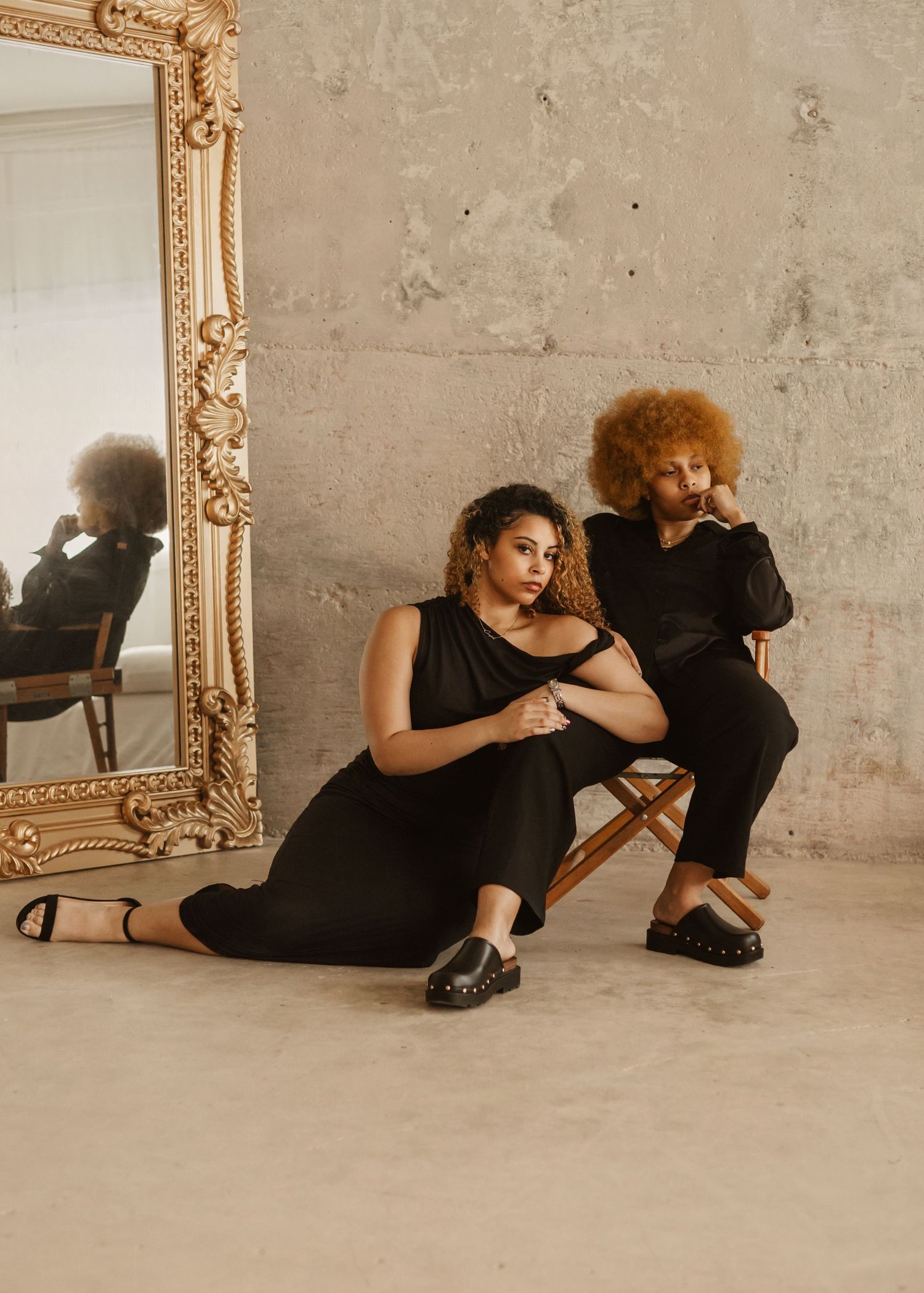 A figure in black clothing poses artistically on the floor beside an ornate gold frame in a minimalist setting.