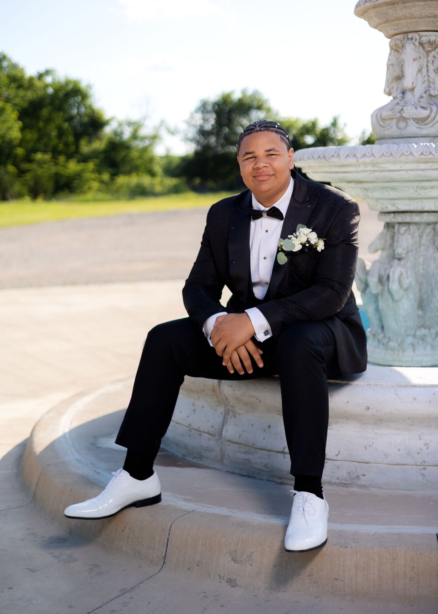Young man in black tuxedo with white boutonniere and white dress shoes sitting by ornate stone fountain.