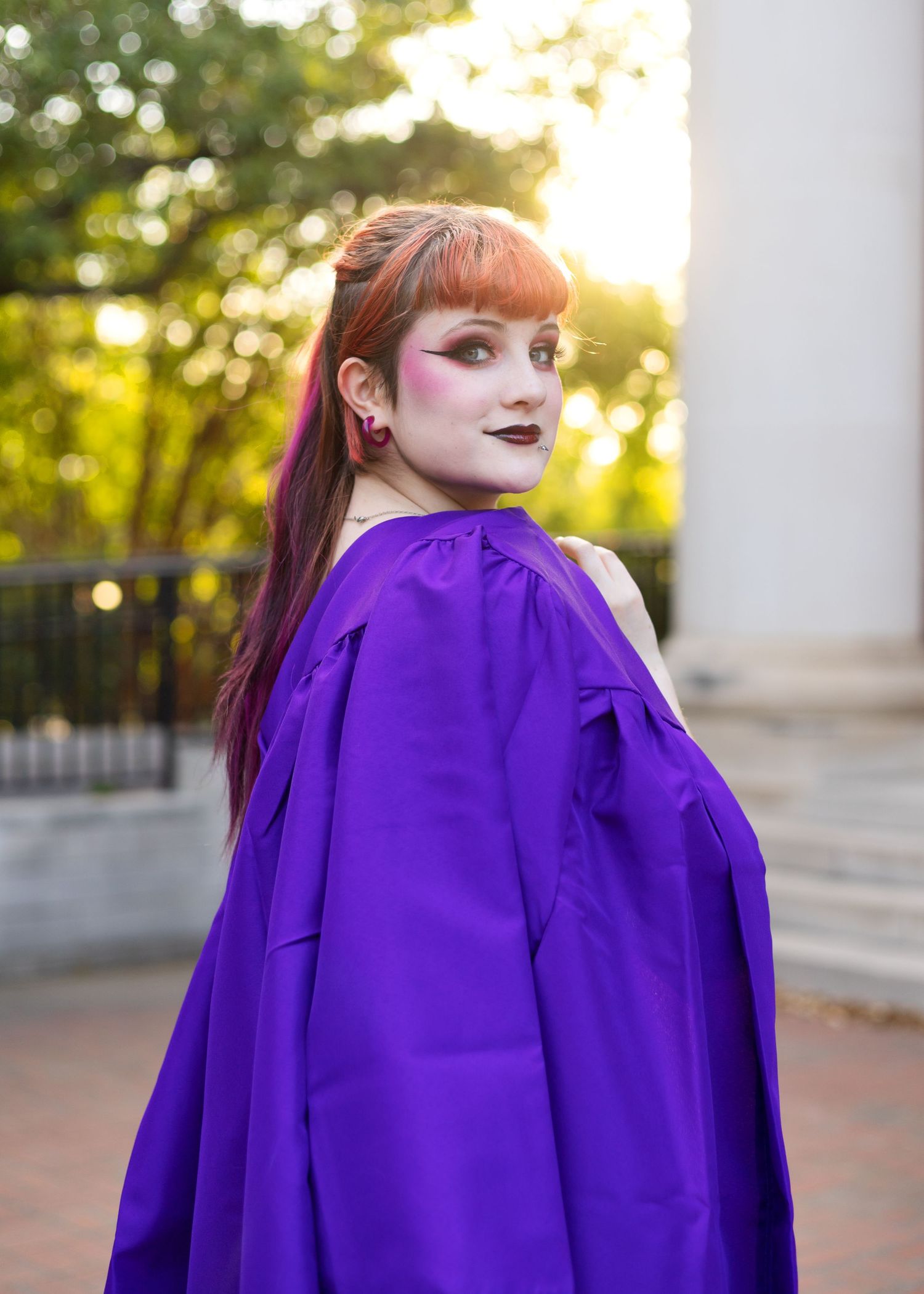 Young woman with red hair wearing a purple graduation gown poses outdoors during golden hour.