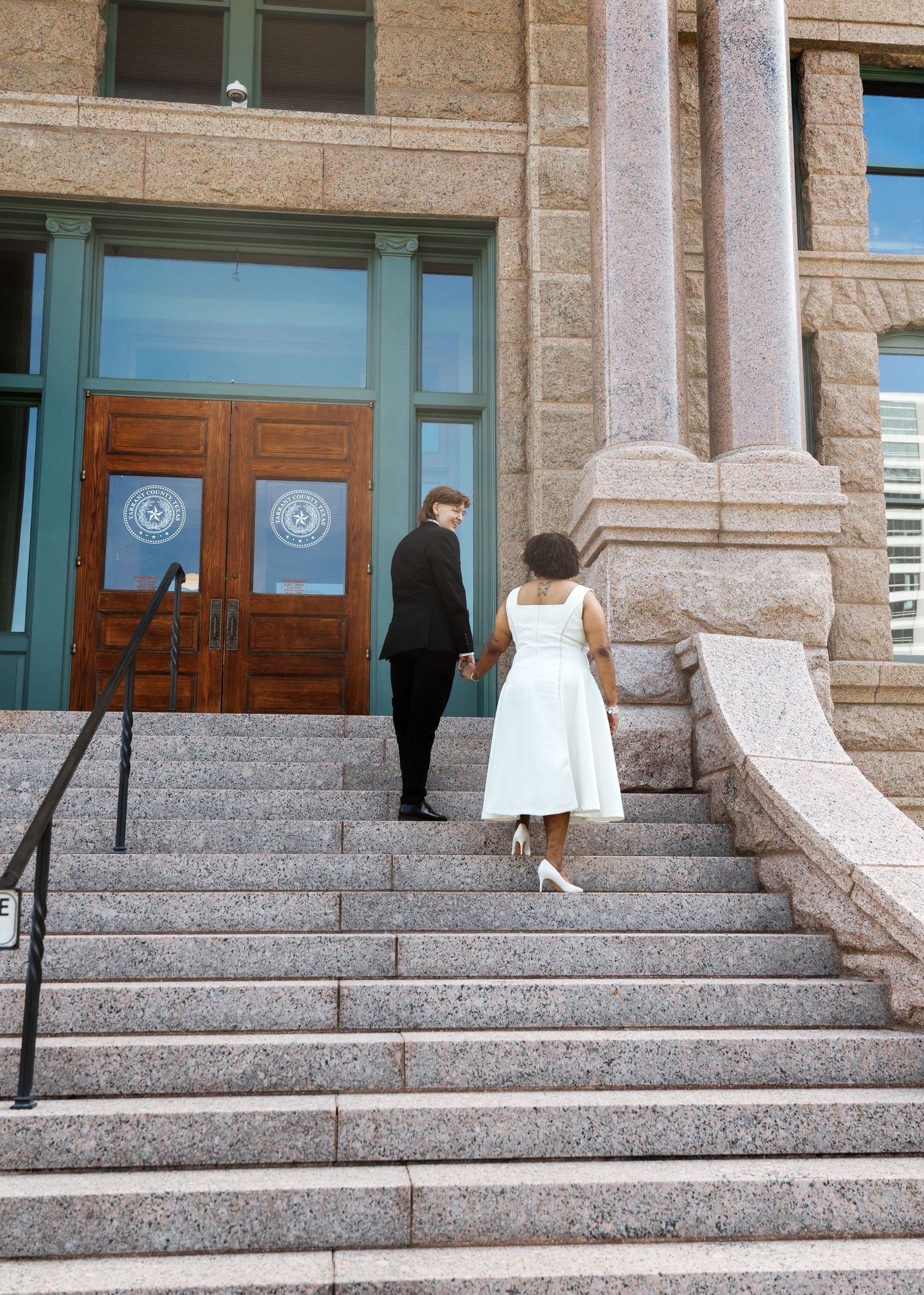 Couple in wedding attire holding hands walking up granite steps toward ornate wooden doors of Texas courthouse.