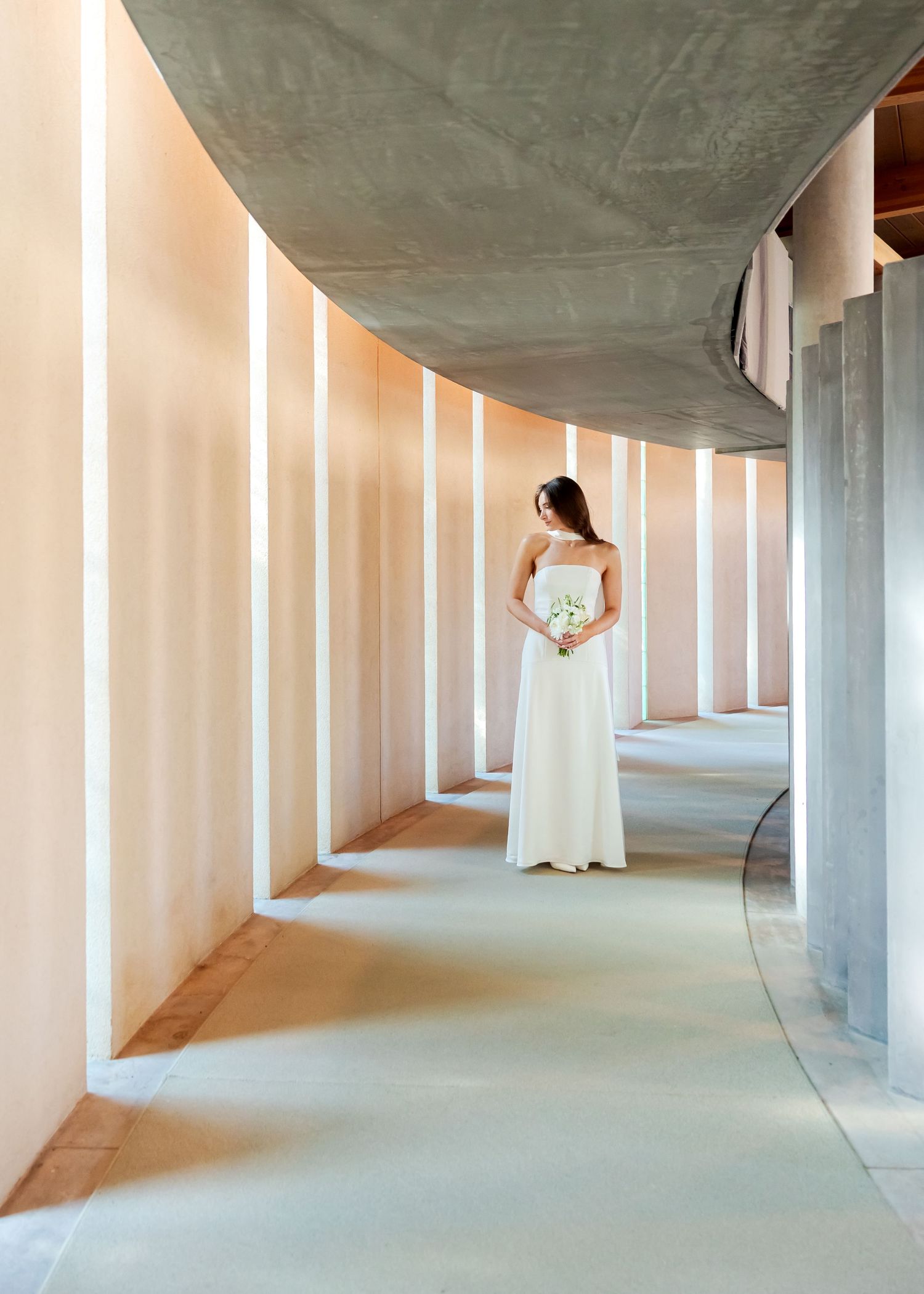 Bride in white gown holding bouquet stands in elegant curved corridor with soft lighting and concrete architecture.