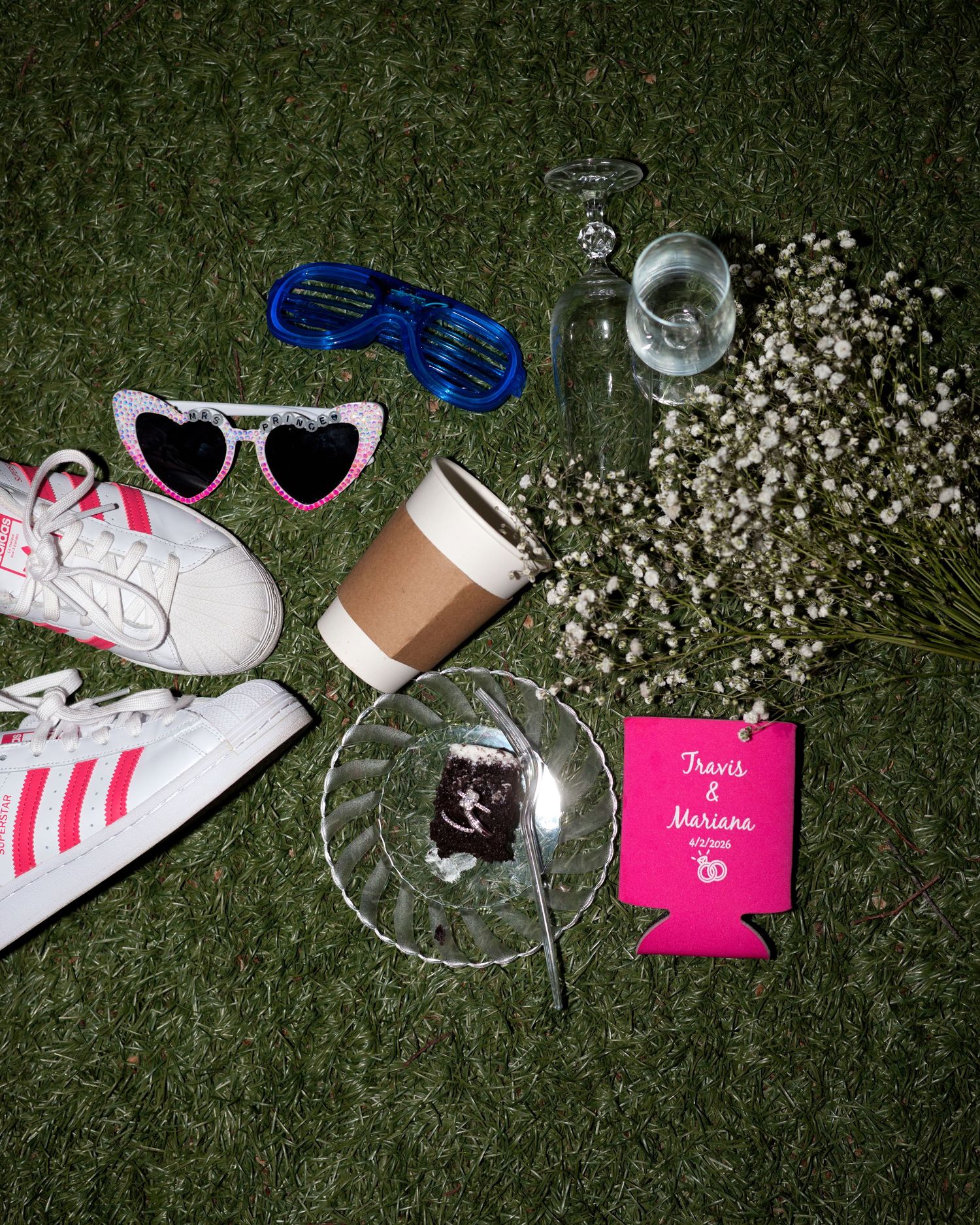 Flat lay of wedding items including Adidas sneakers, heart sunglasses, coffee cup, flowers, and pink Travis & Mariana koozie on grass.
