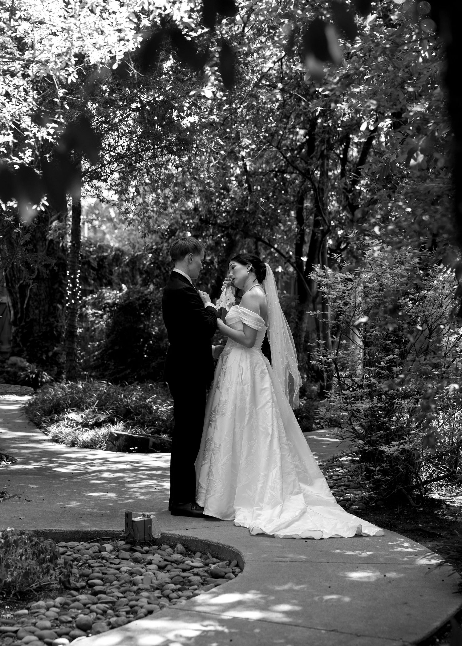 Black and white photo of a bride and groom holding hands on a garden path surrounded by lush trees.