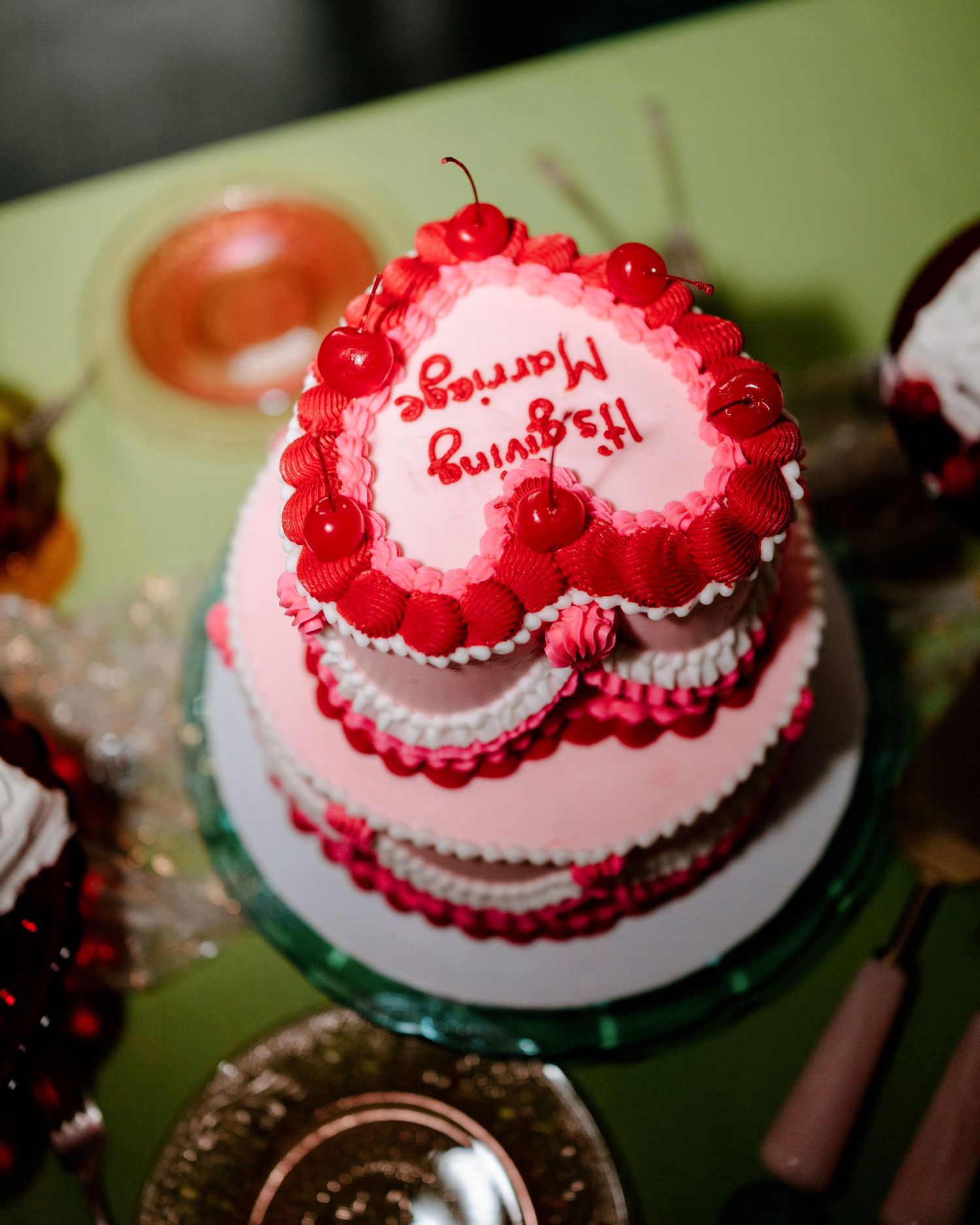 Heart-shaped pink wedding cake with red cherries and Happy Marriage inscription on green table.
