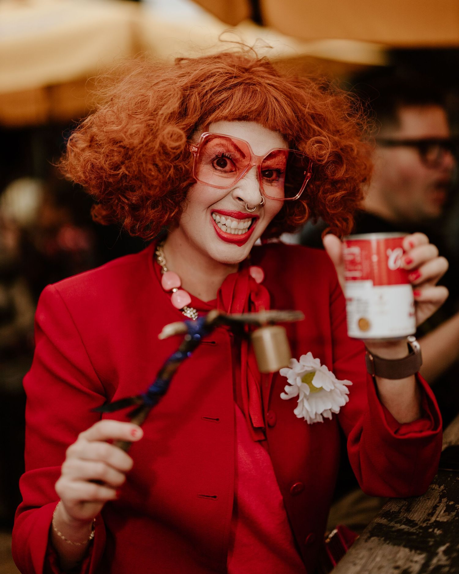 A person with curly red hair and bright red lipstick wearing a red sweater smiles while holding up an ID card.