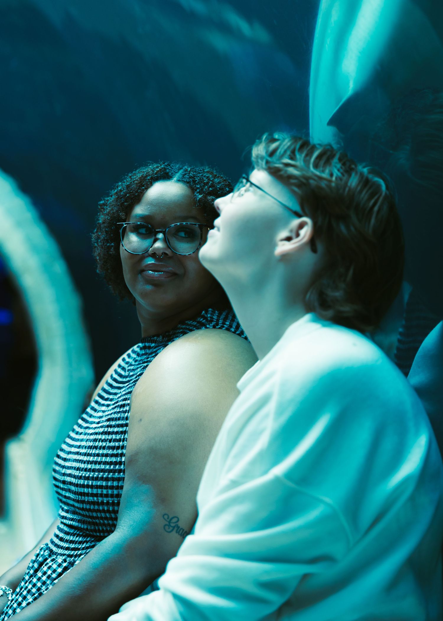 Two people sharing a tender moment in teal-lit venue, woman with glasses smiling warmly beside companion.