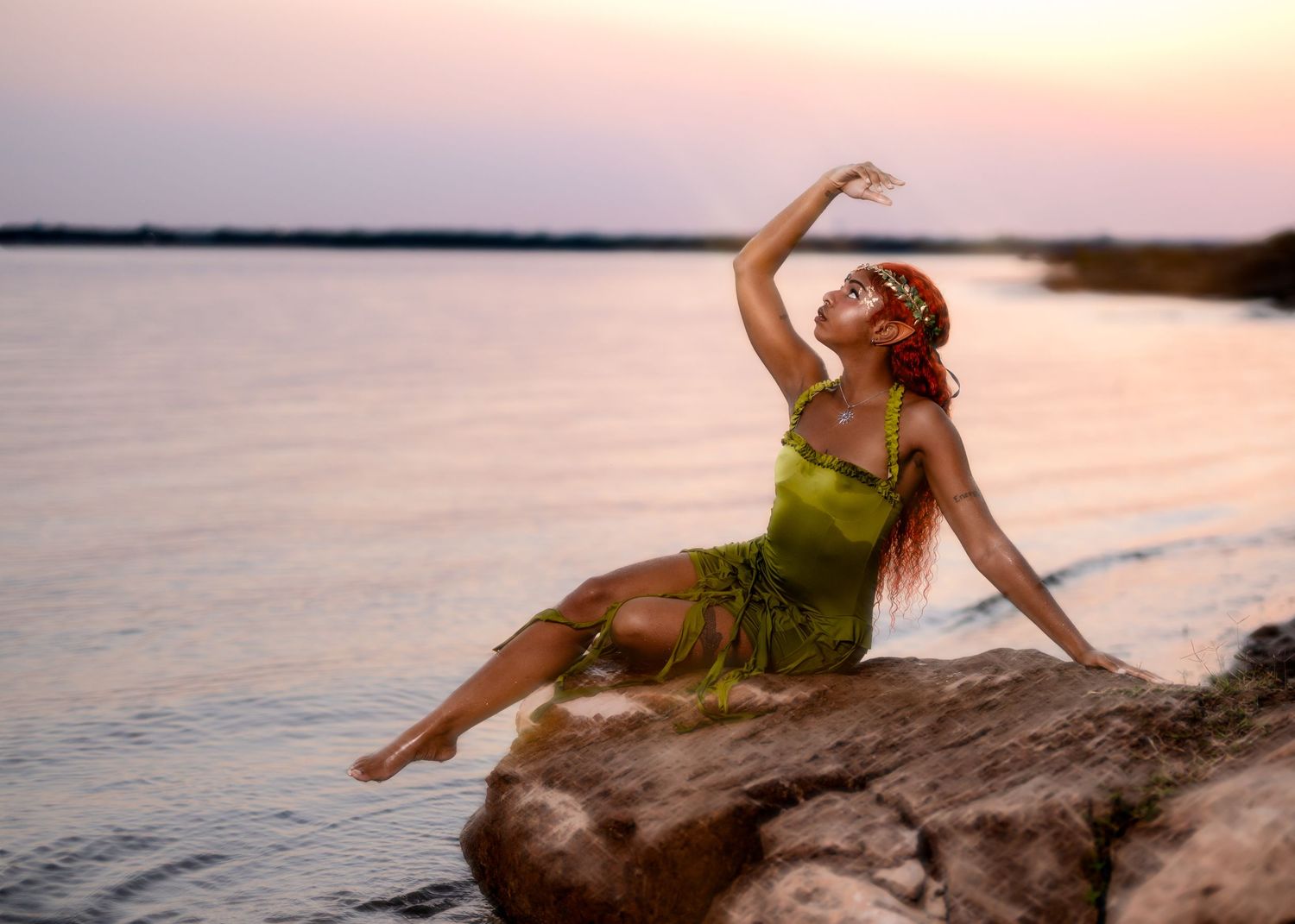 A person in a green dress poses gracefully on coastal rocks during a pink sunset over the ocean.