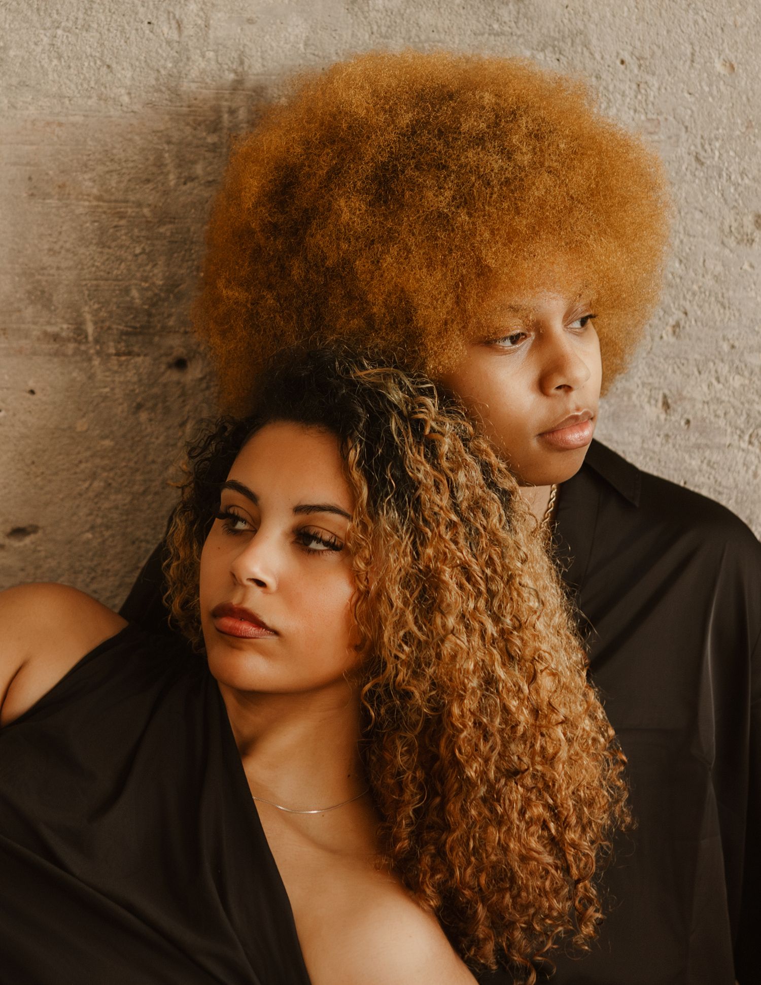 Portrait of a person with vibrant copper-colored curly hair against a textured wall in warm lighting.