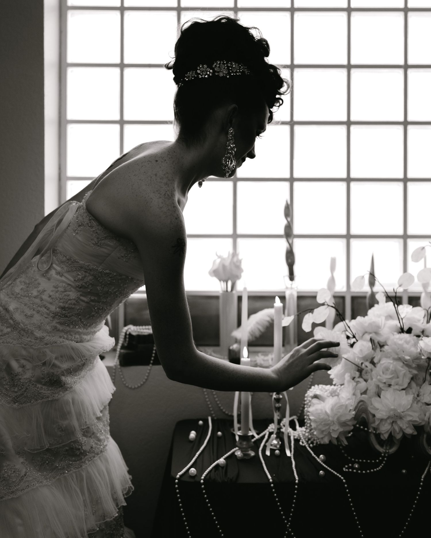 Black and white photo of a bride in a wedding dress arranging white floral centerpiece near a window.