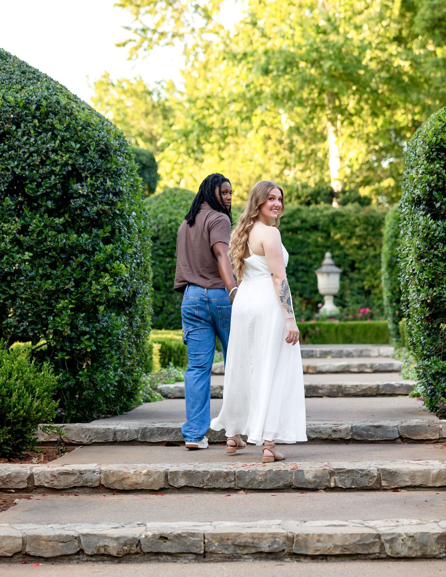 Couple in formal wear walking through a lush garden pathway with trimmed hedges and stone steps.