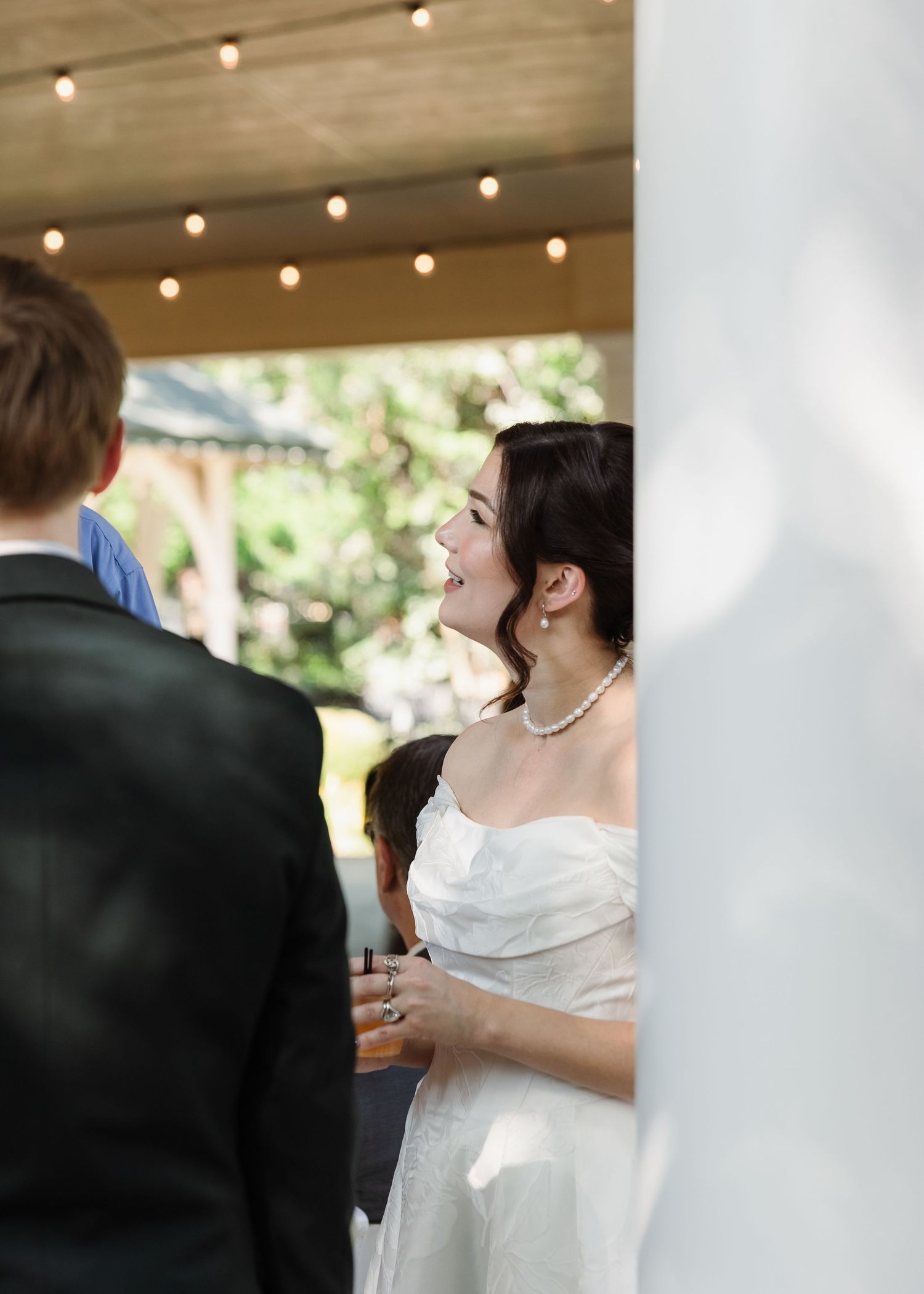 Bride in white off-shoulder gown and pearl necklace laughing joyfully with groom at outdoor wedding reception.