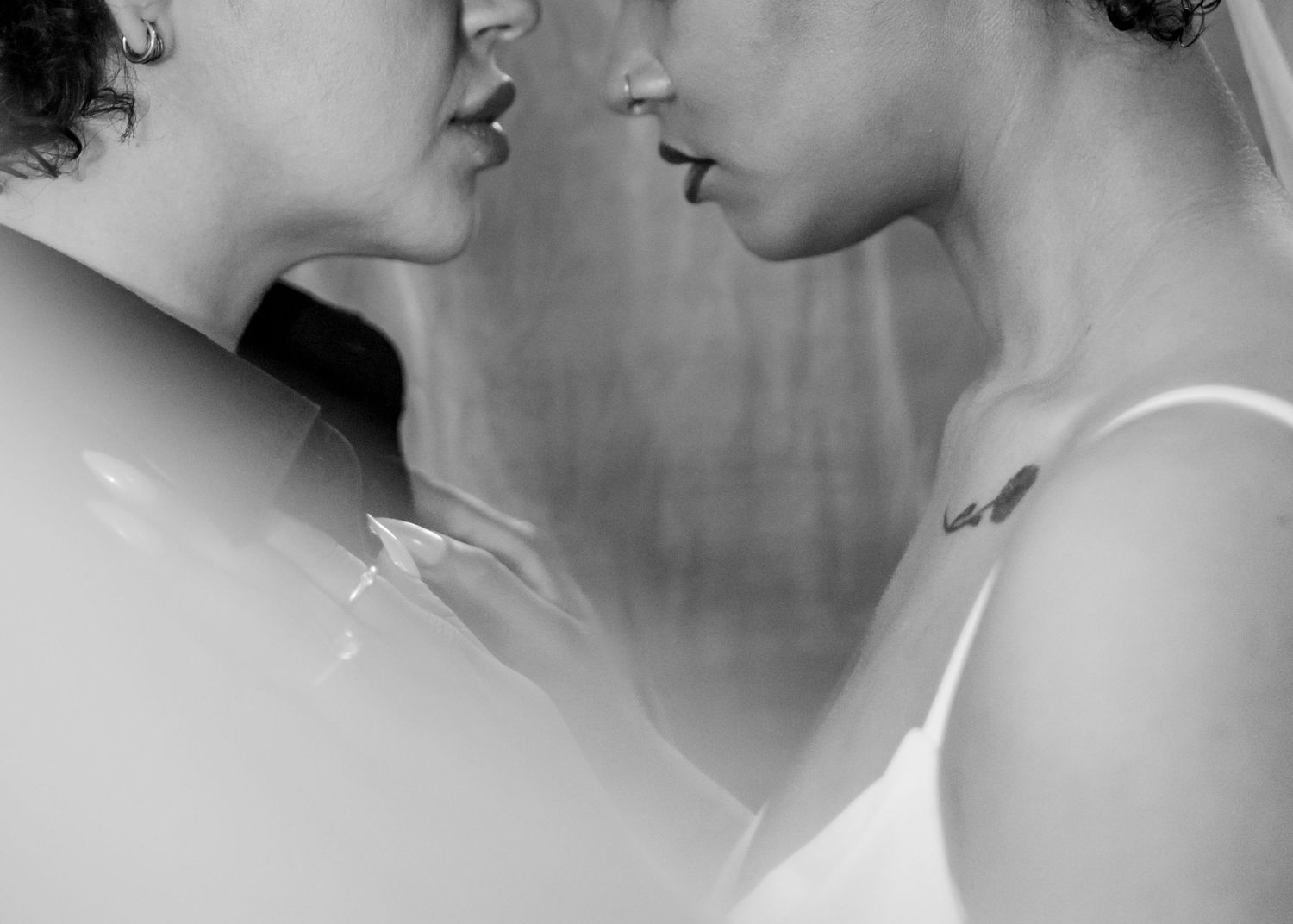 Black and white photo of two women facing each other intimately, one with a small tattoo on her neck.