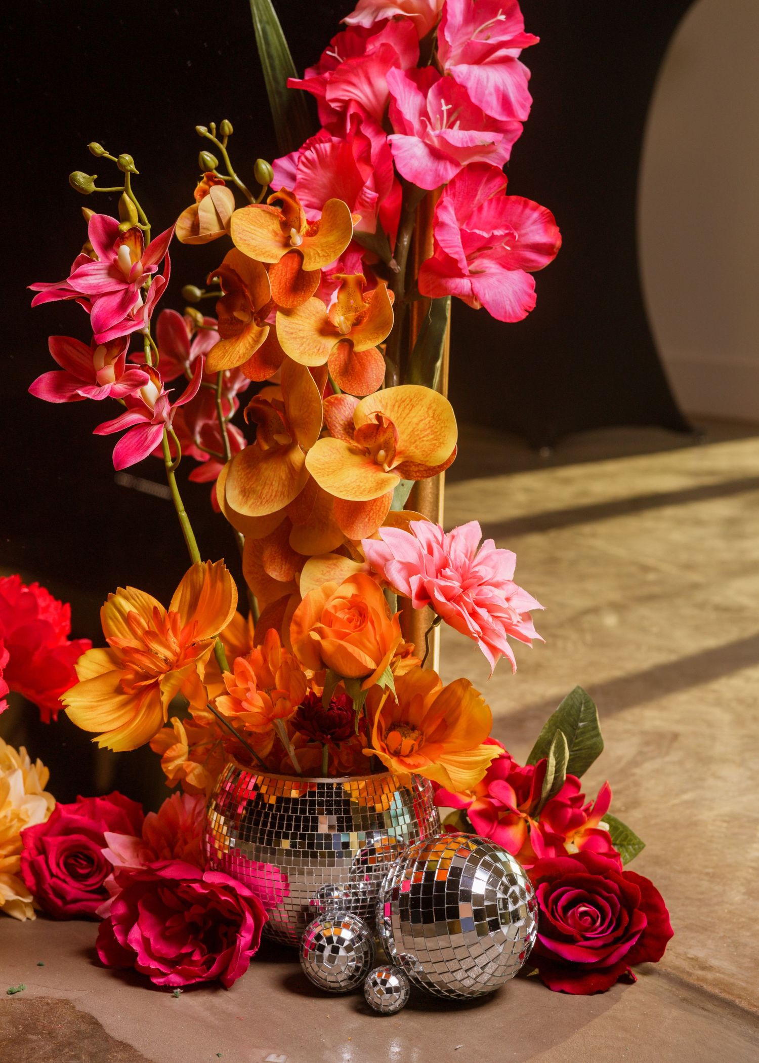 Vibrant floral arrangement with pink, orange, and red flowers surrounding silver disco balls on a marble surface.
