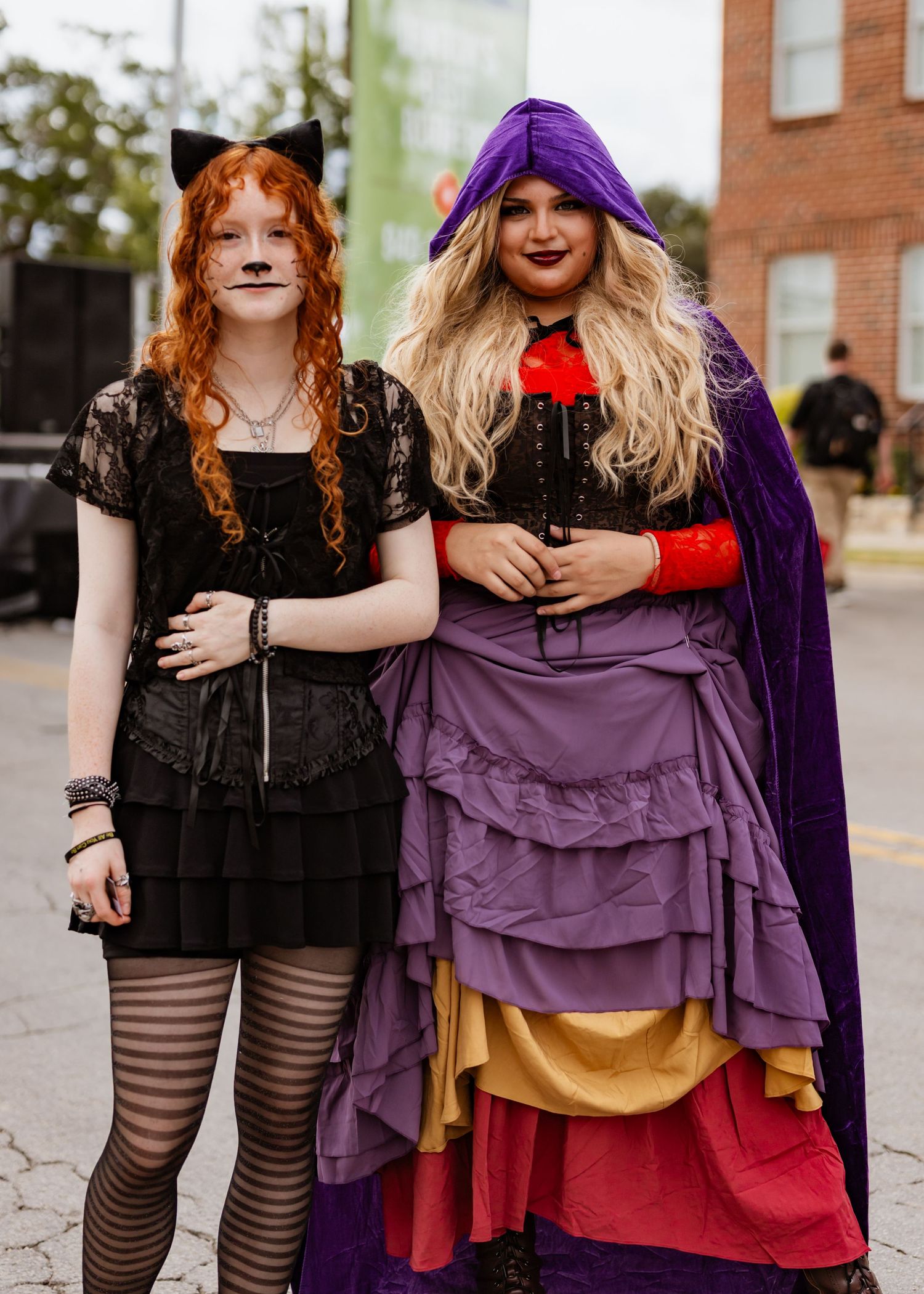 Two people in gothic Halloween costumes with striped stockings and purple dress pose together outdoors.