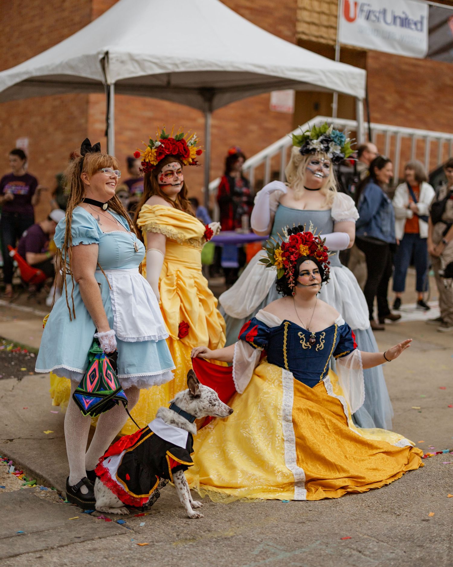 People in colorful princess-style ball gowns gather at an outdoor festival with tents in the background.