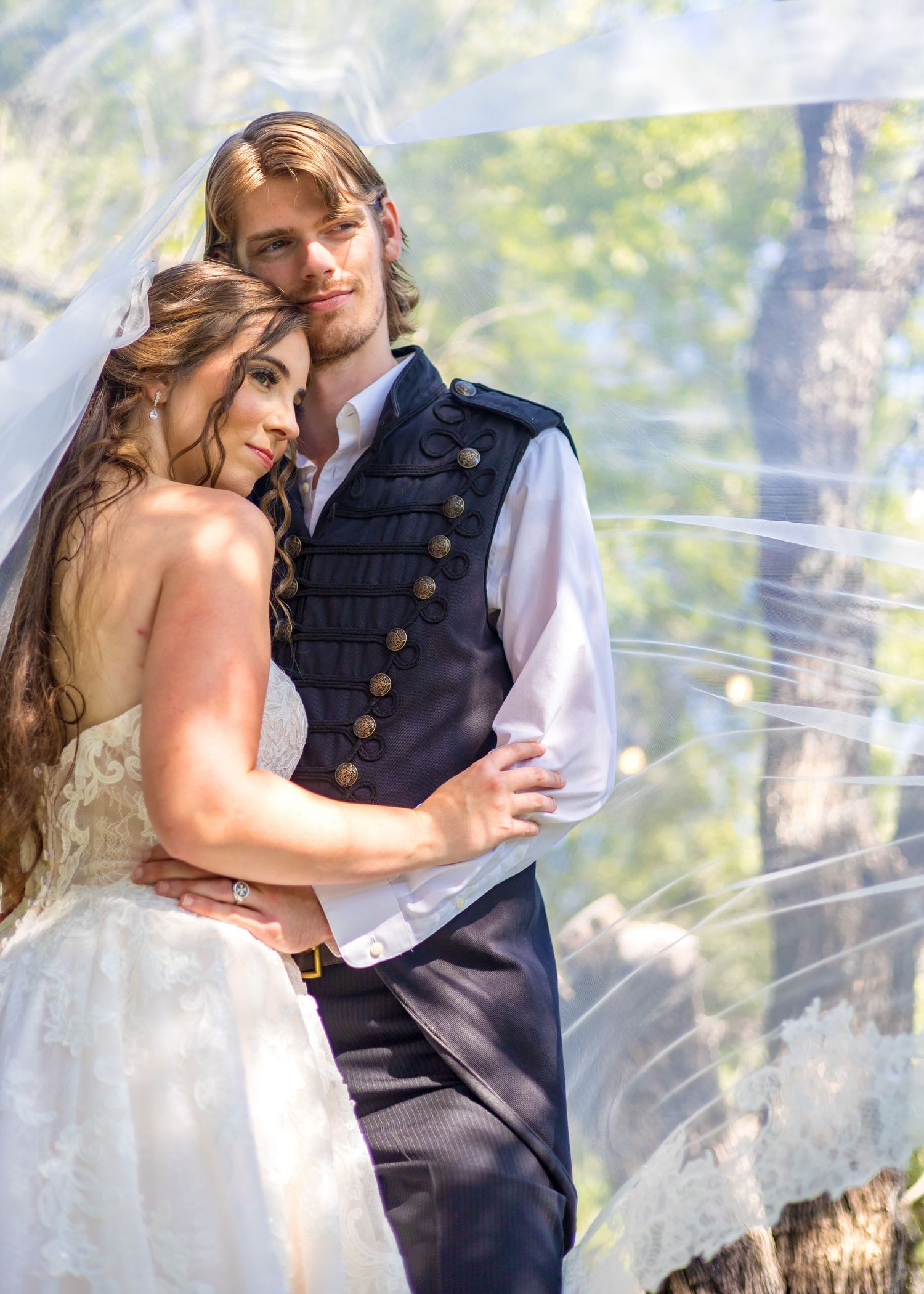 Bride and groom in elegant wedding attire, groom wearing military-style vest, posing romantically outdoors under a flowing veil.