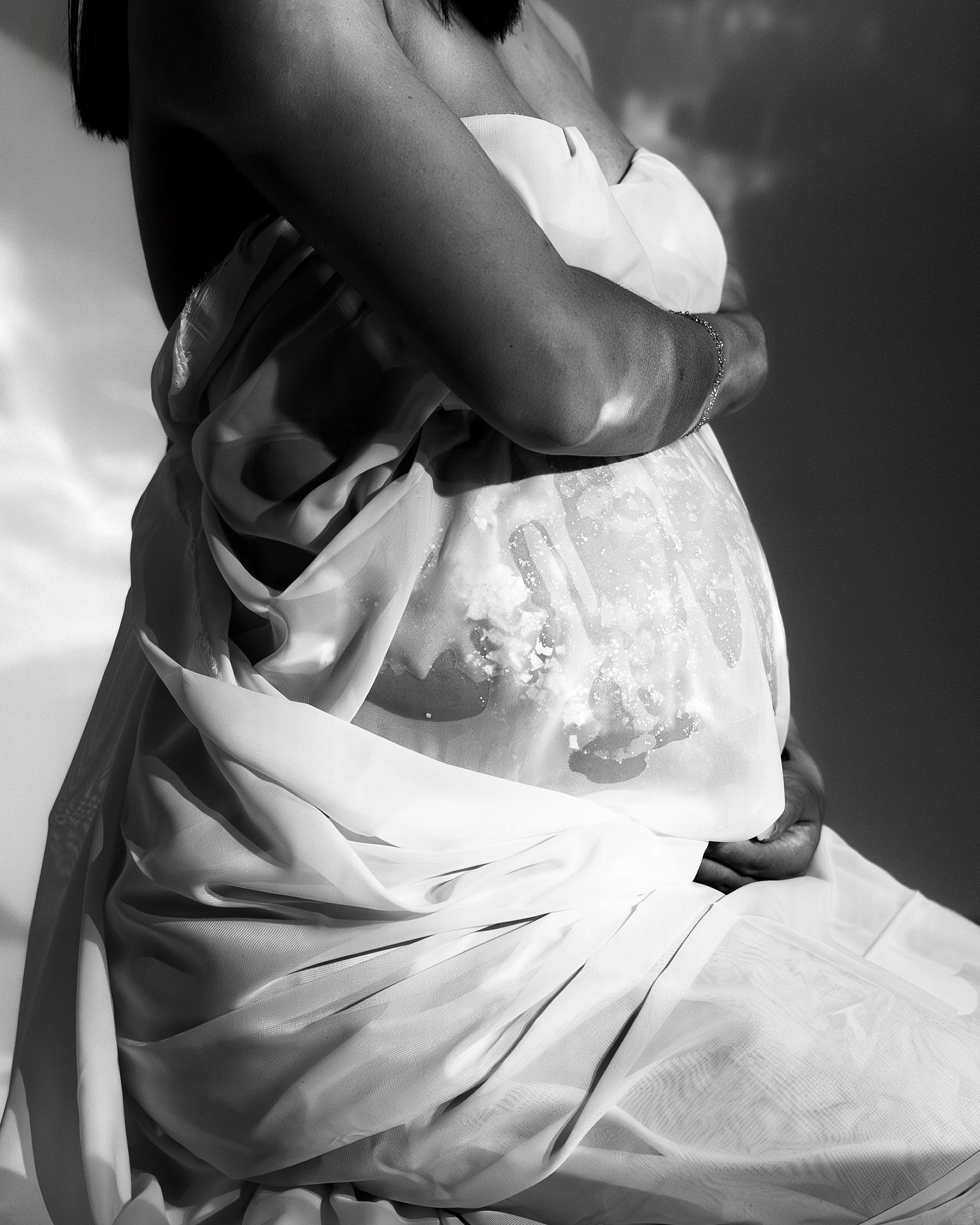 Black and white maternity photo of pregnant woman cradling baby bump draped in sheer white fabric.