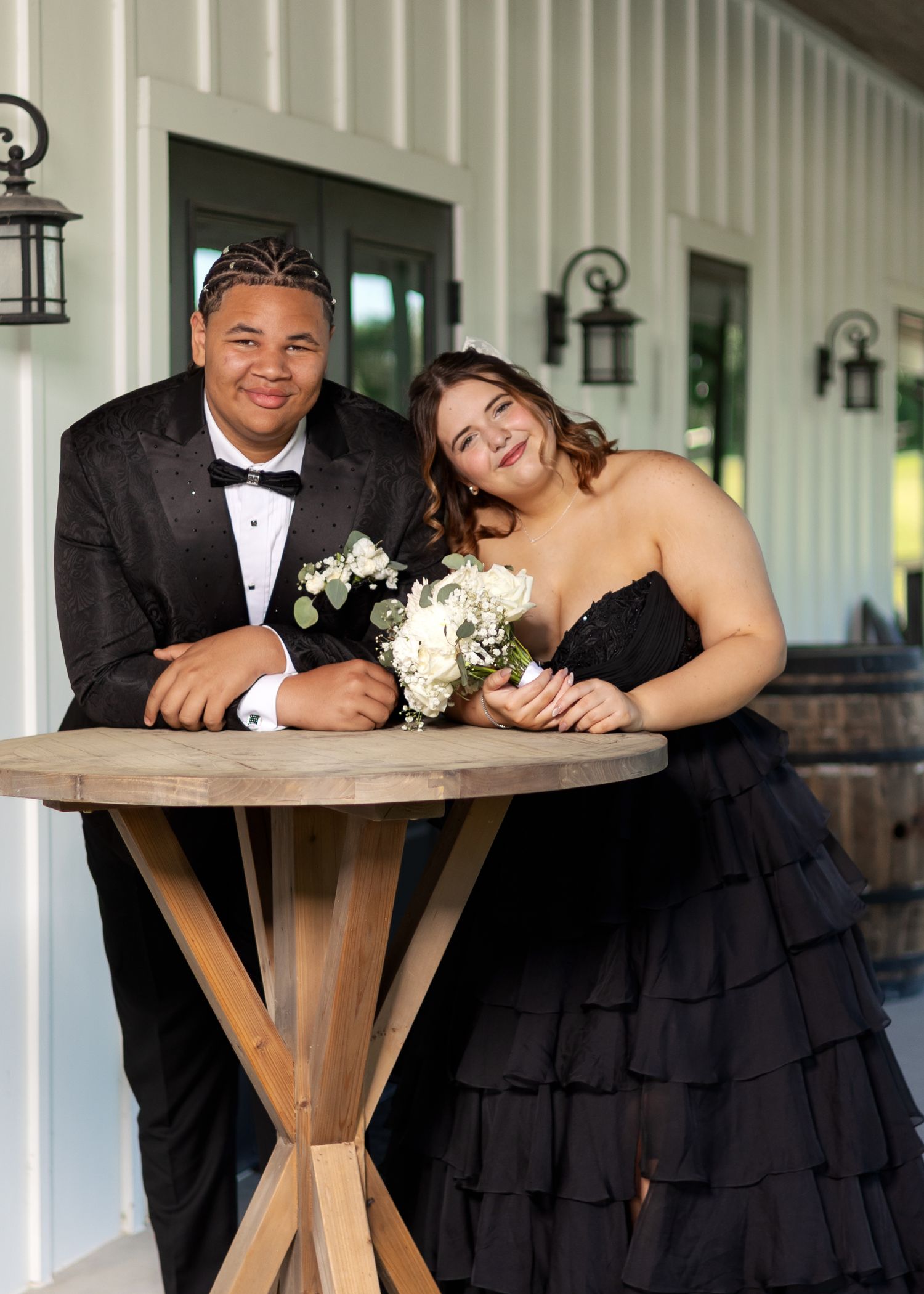 Couple in black formalwear posing at a wooden table, man in tuxedo and woman in strapless gown holding white bouquet.