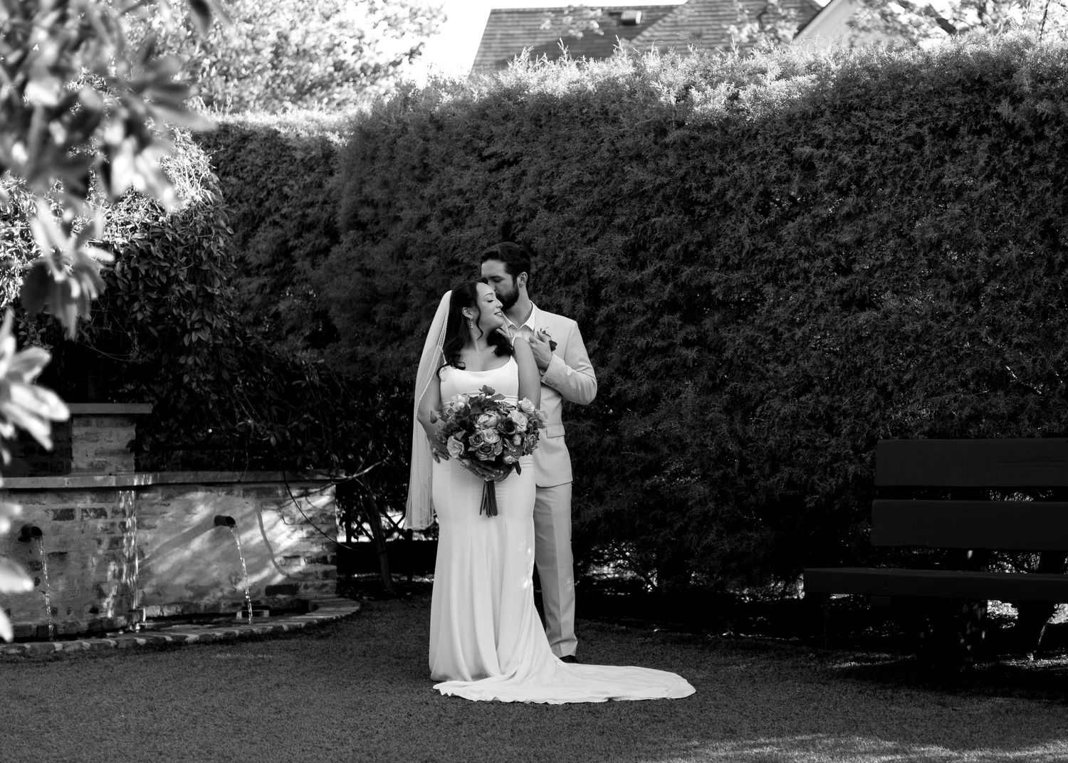 Black and white wedding photo of bride and groom embracing in a garden with lush hedges backdrop.