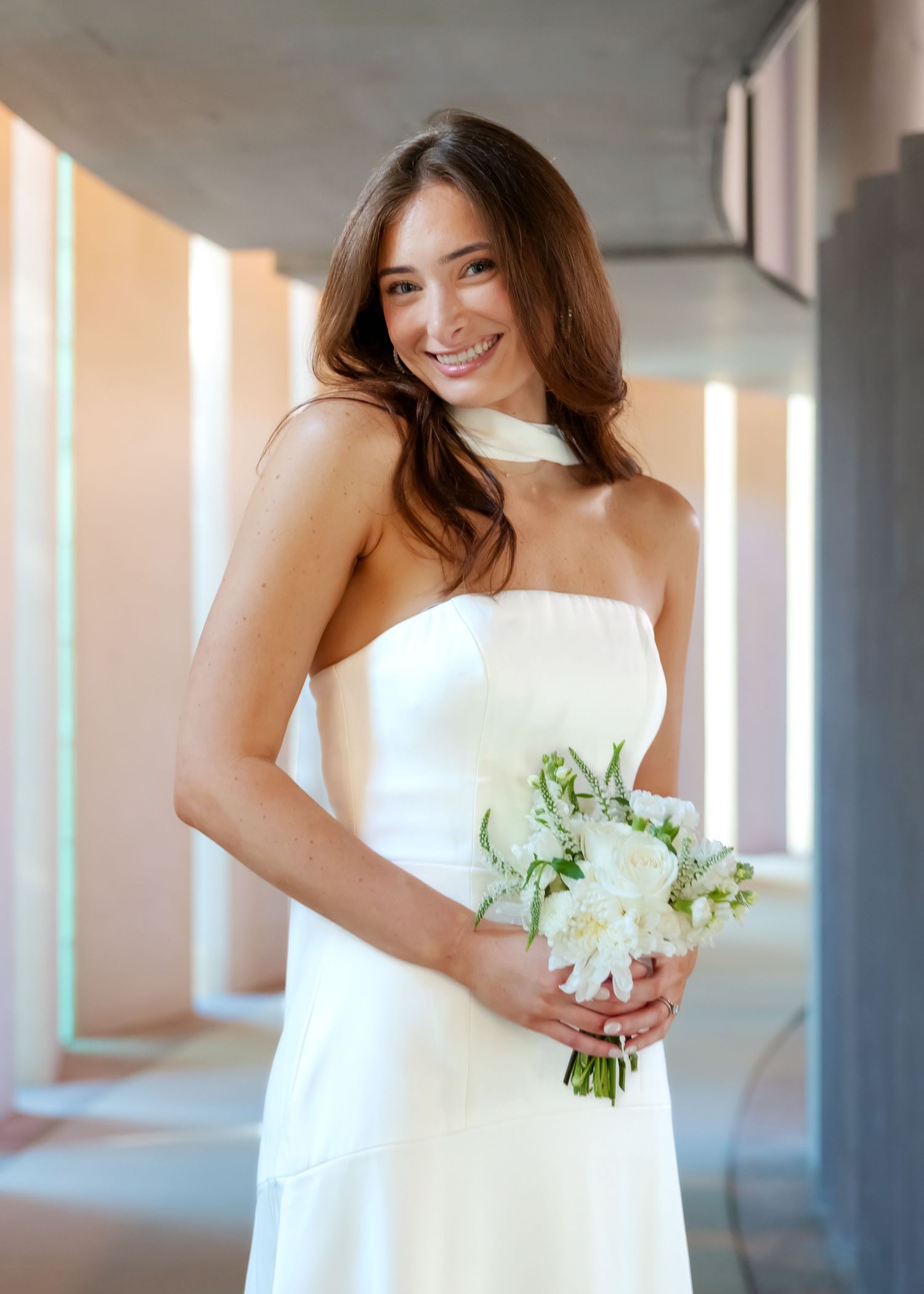Smiling bride in strapless white gown holding white flower bouquet in modern architectural hallway.