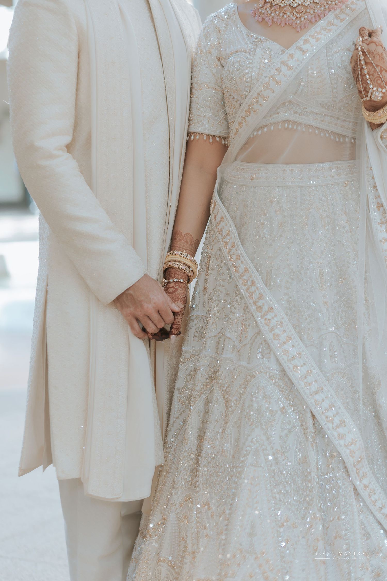 Indian bride and groom in matching white outfits holding hands, showcasing bridal bangles and lehenga.