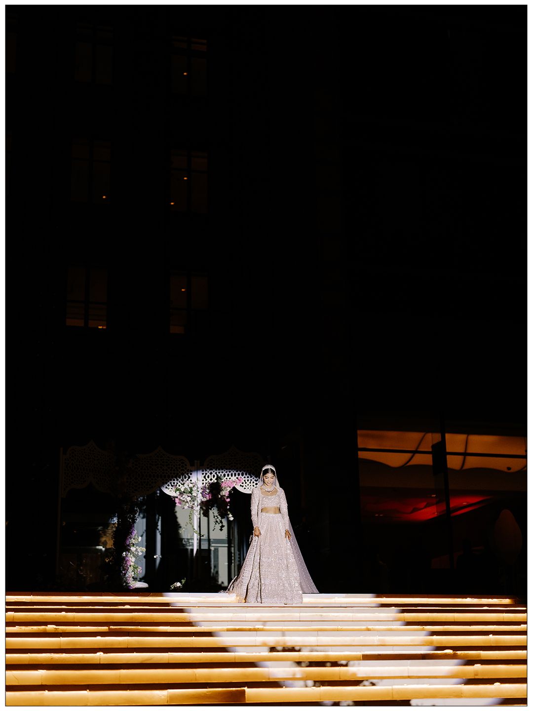 Bride in embroidered lehenga and veil stands illuminated on grand staircase during nighttime wedding ceremony.