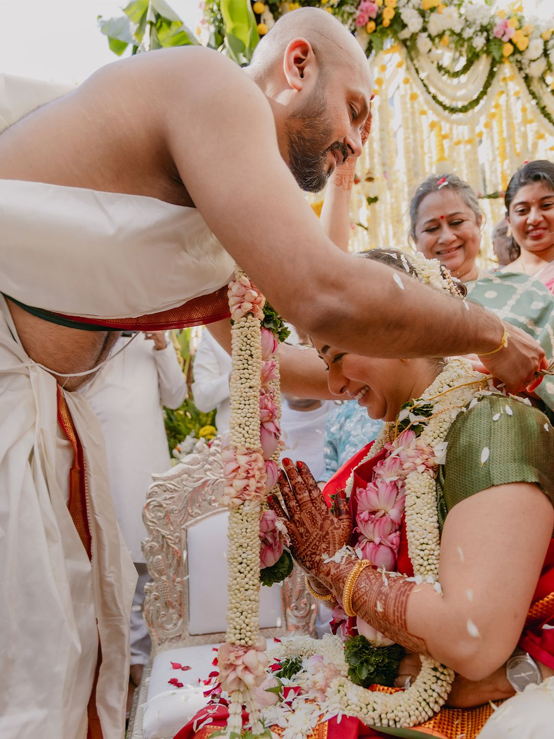 Bald groom places garland on bride during traditional South Indian wedding ceremony with smiling guests in background.