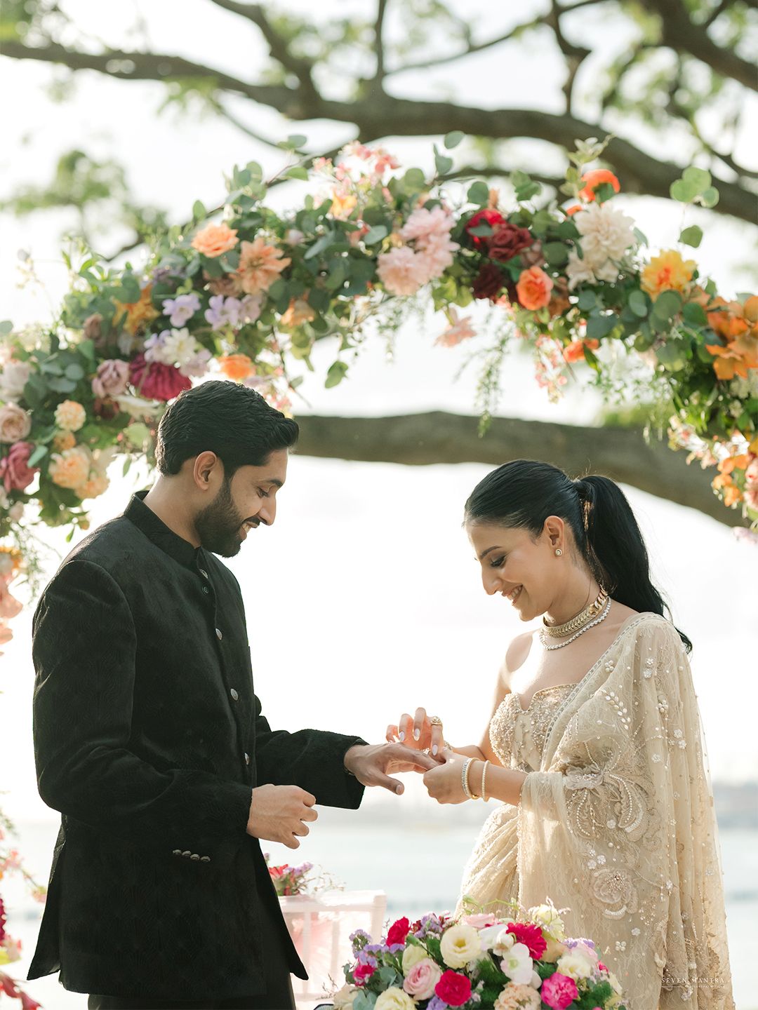 Groom in black sherwani placing ring on brides finger under a colorful floral wedding arch.