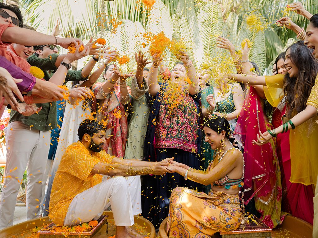 Joyful Haldi ceremony with couple showered in flower petals by colorfully dressed wedding guests outdoors.