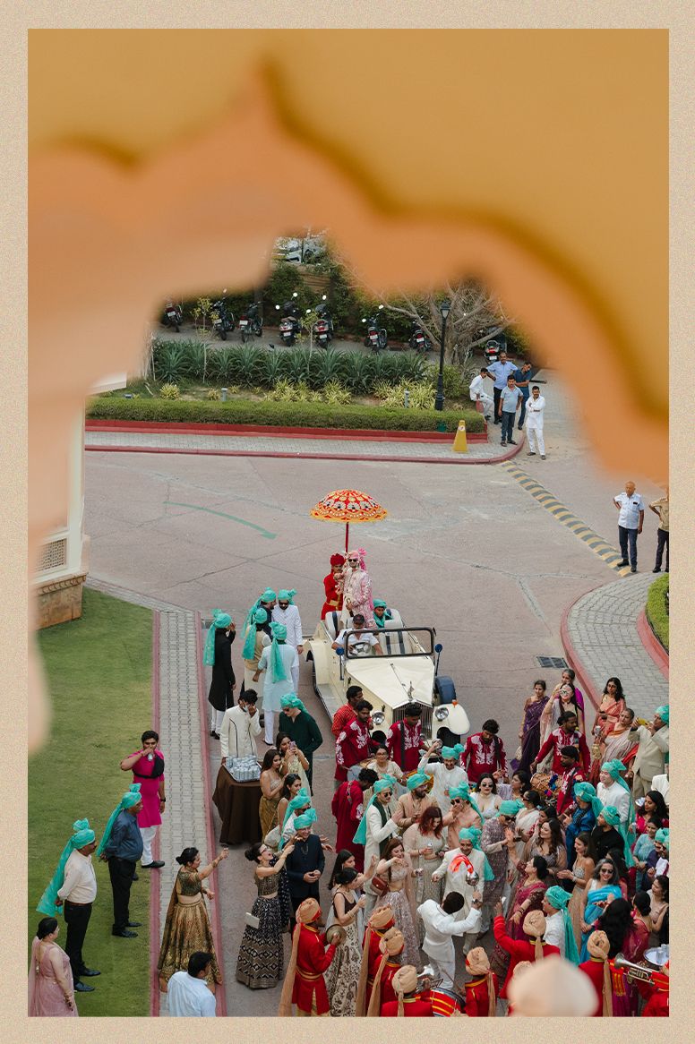Aerial view of colorful Indian baraat procession with orange umbrella and guests in traditional attire.