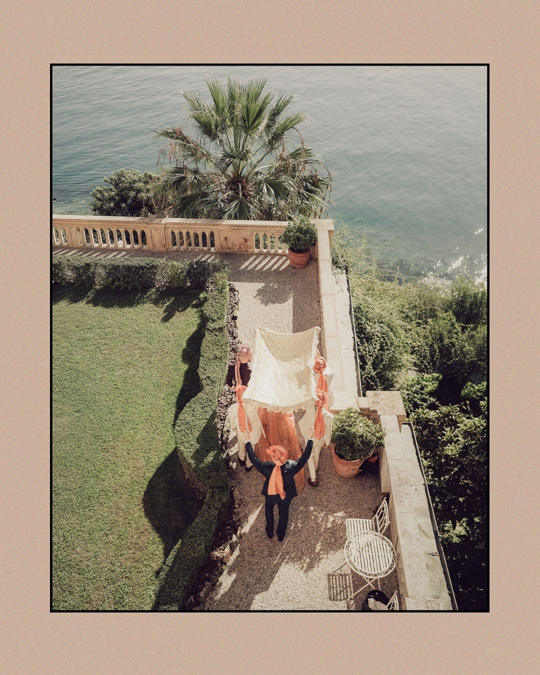 Aerial view of Indian wedding couple walking under floral canopy on lakeside villa terrace.