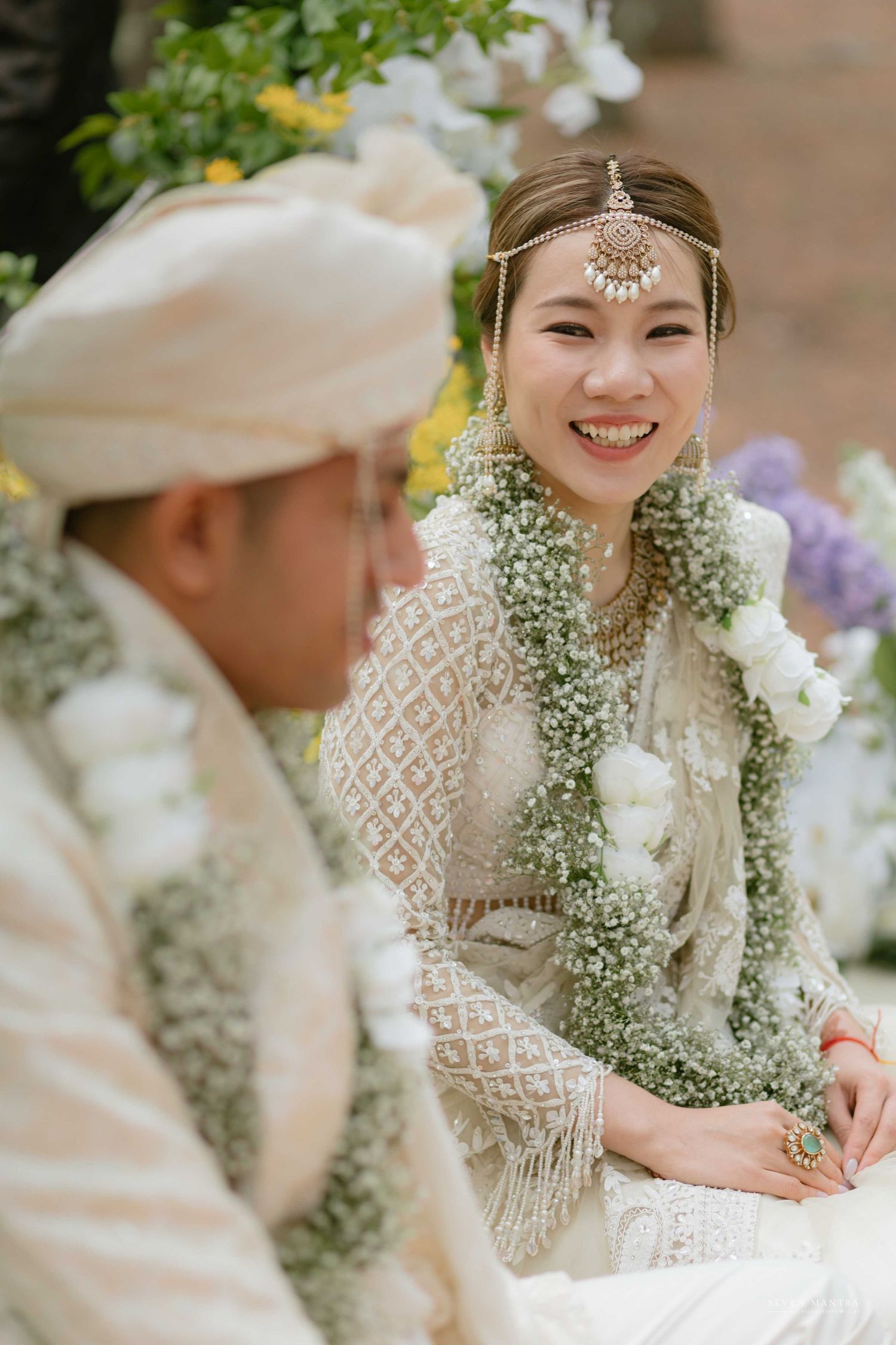 Smiling bride in white lace saree adorned with floral garlands during Hindu wedding ceremony.