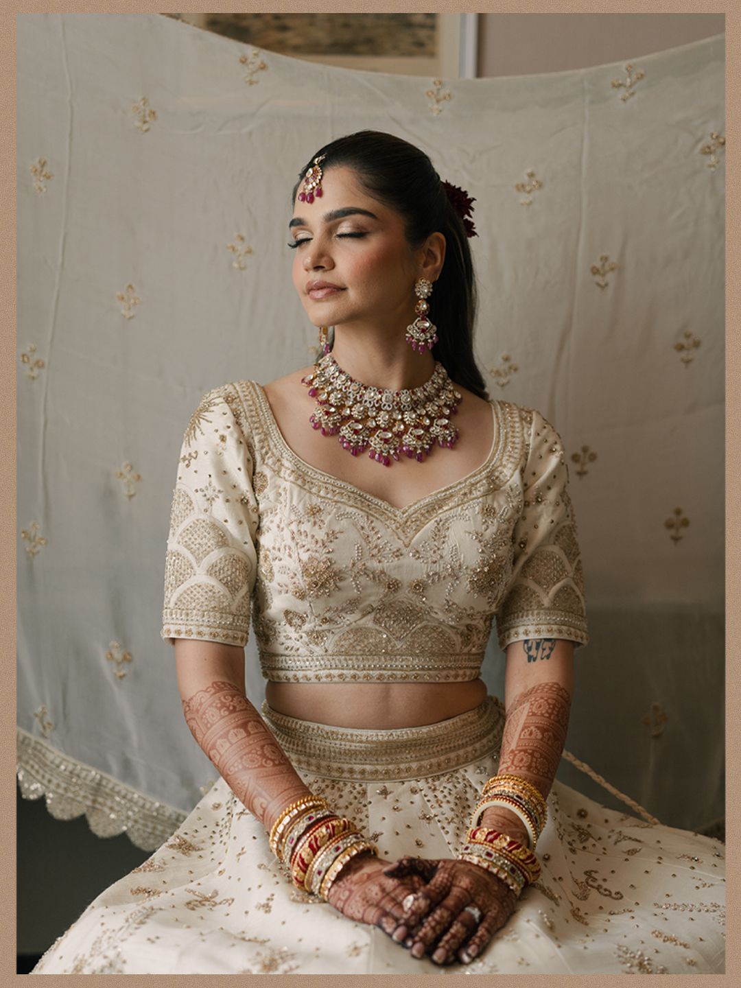 Indian bride in cream lehenga with ruby and pearl necklace and mehndi hands in soft light portrait.
